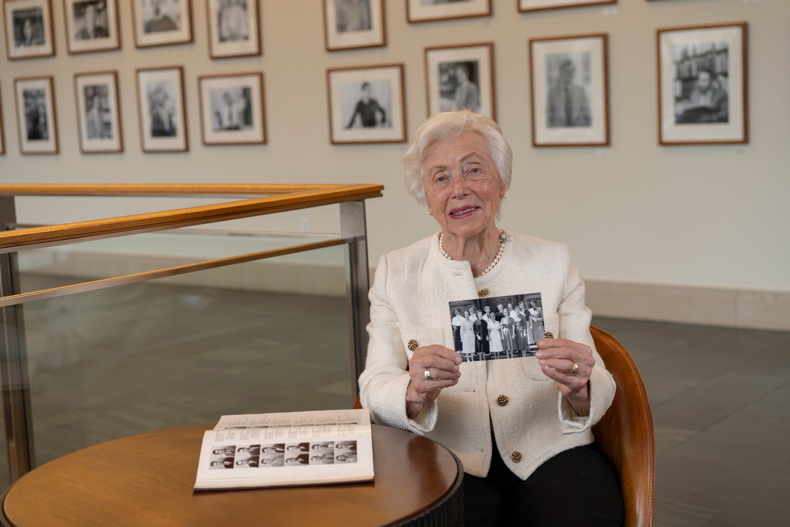 A woman wearing a white jacket sits at a table on which a book is opened to pages of headshots. She holds a photograph of a group of women posing on the steps of a building.