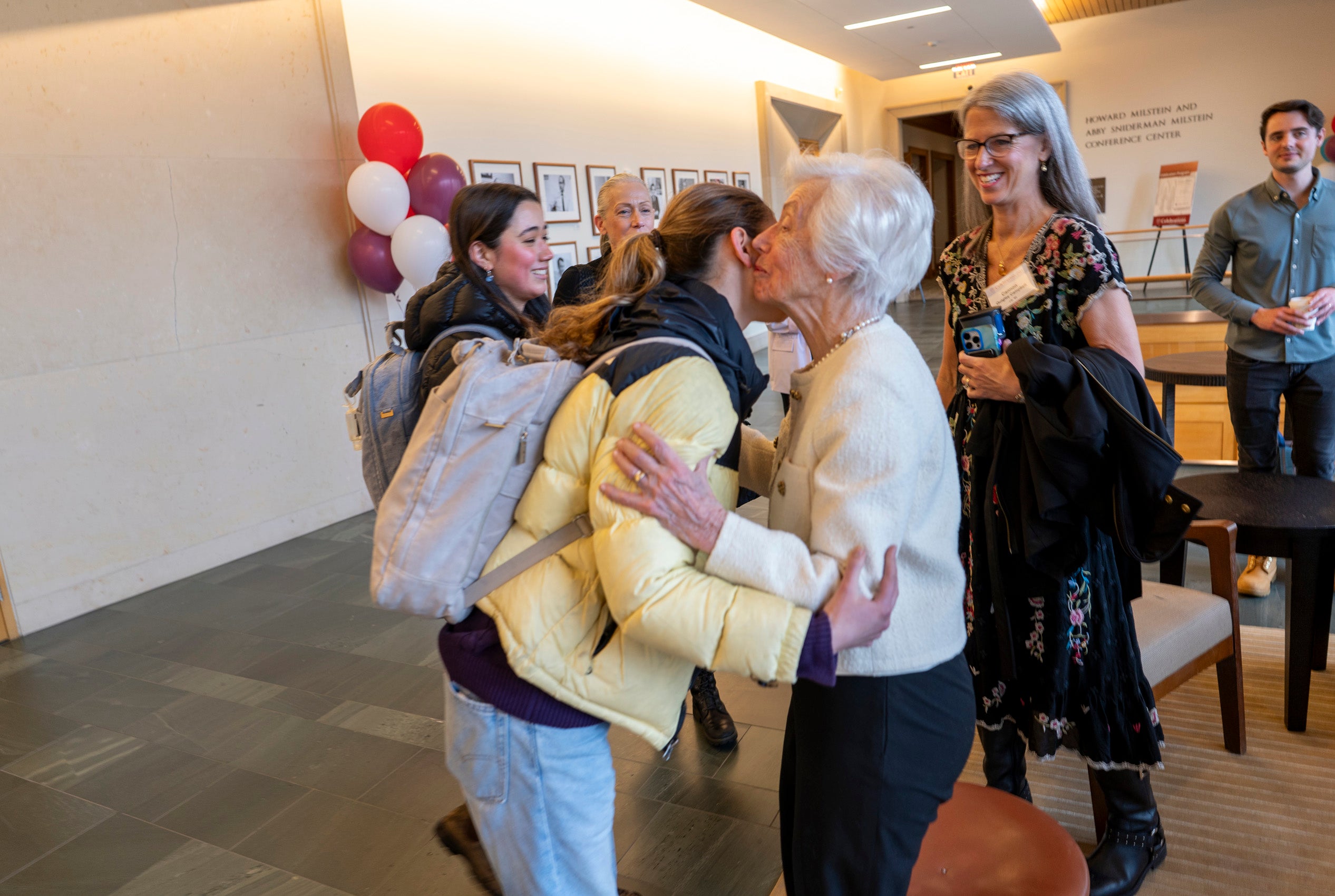 A young woman greets an older woman as others look on.