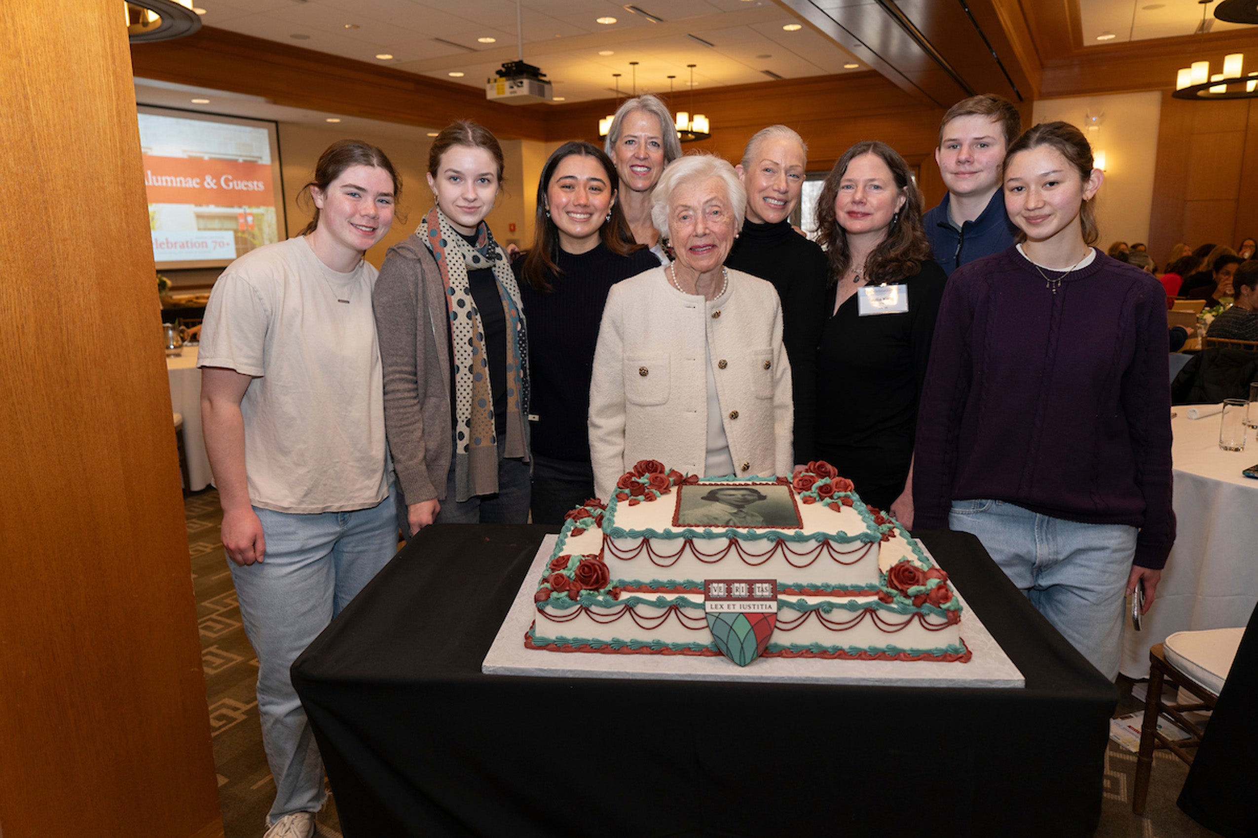 A group portrait of 9 family members around a birthday cake in a large room.