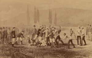 Two groups of men some with dark striped shirts and some with light colored shirts in field playing a sport as onlookers observer