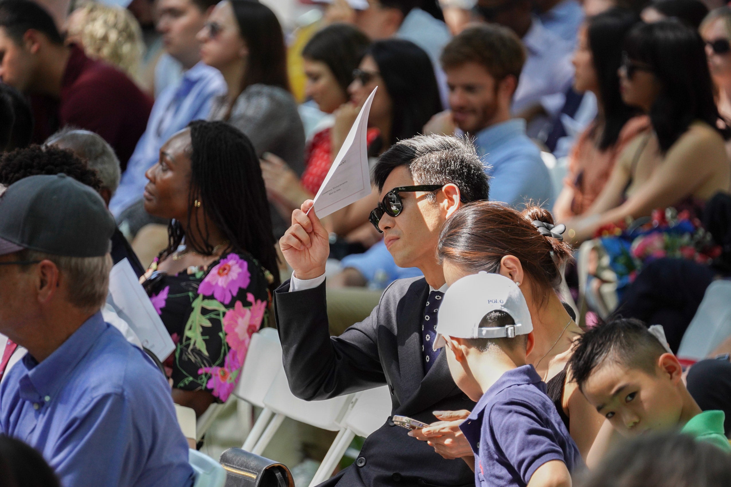 a man holds a program over his eyes for shade as he sits in a crowd watching an event.