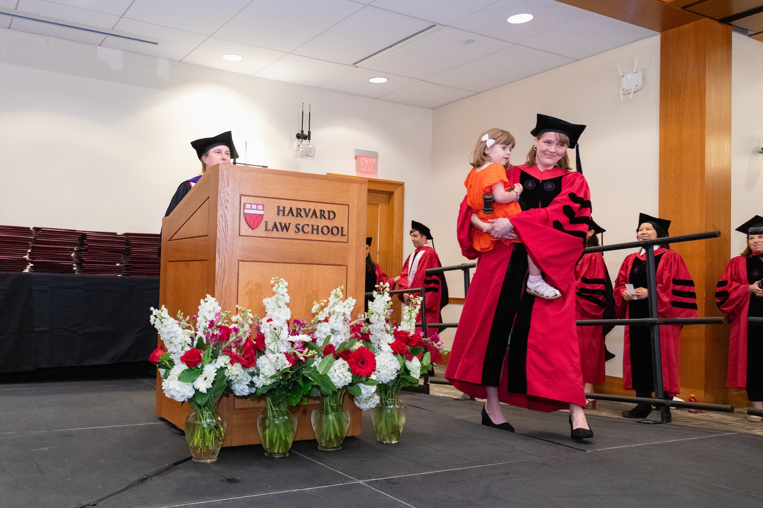 a woman in a red gown and black cap walks across a stage carrying a child.