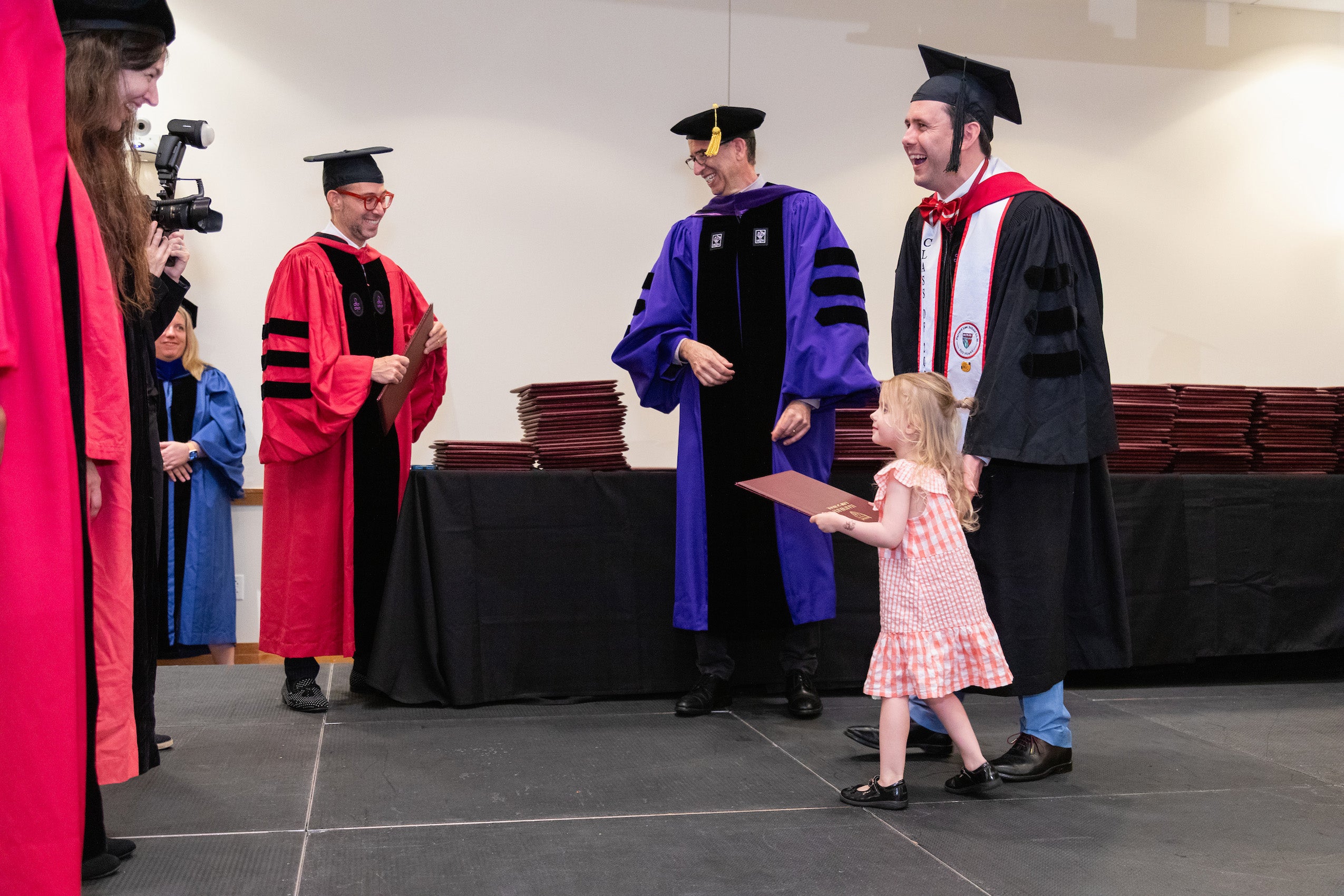 A man in a cap and gown smiles with his young daughter holding his diploma goes across the stage
