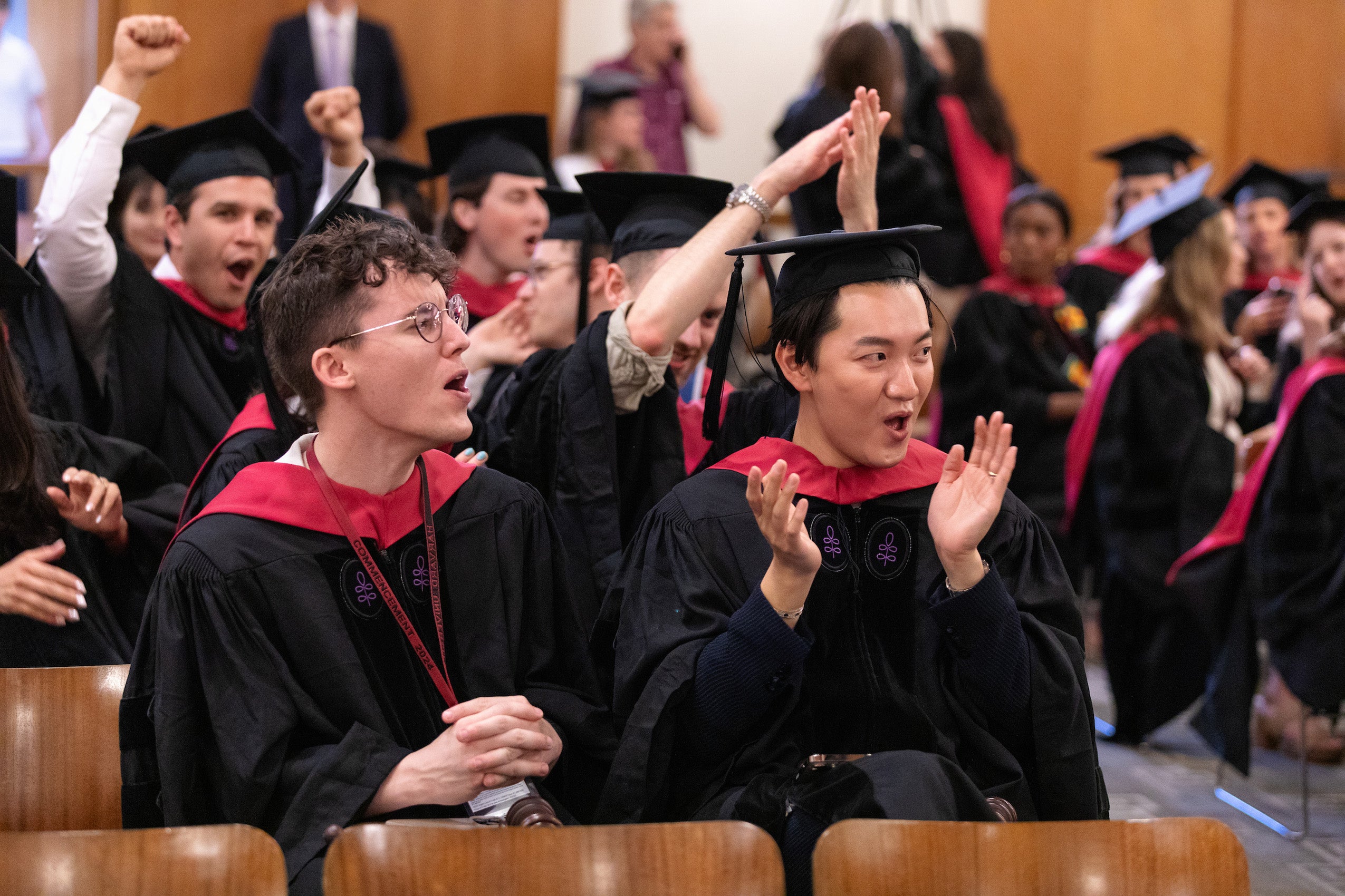 Two men in cap and gown cheer and clap with others in the room from their seats at an event