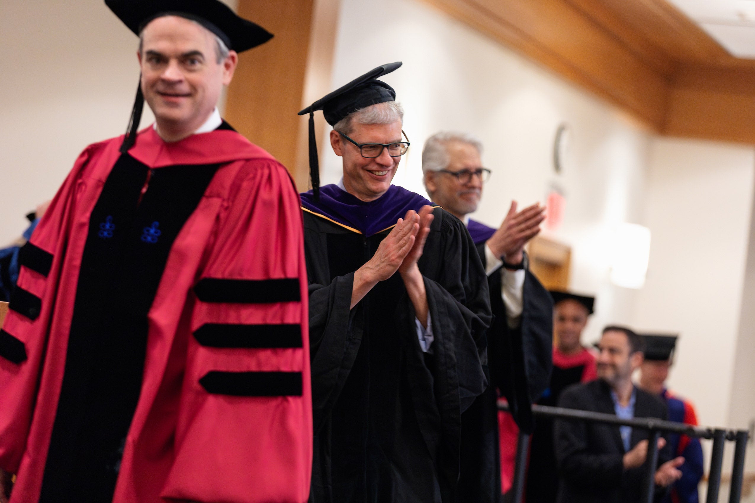 a man in cap and gown claps as he goes across the stage as part of a line of faculty during commencement exercises.