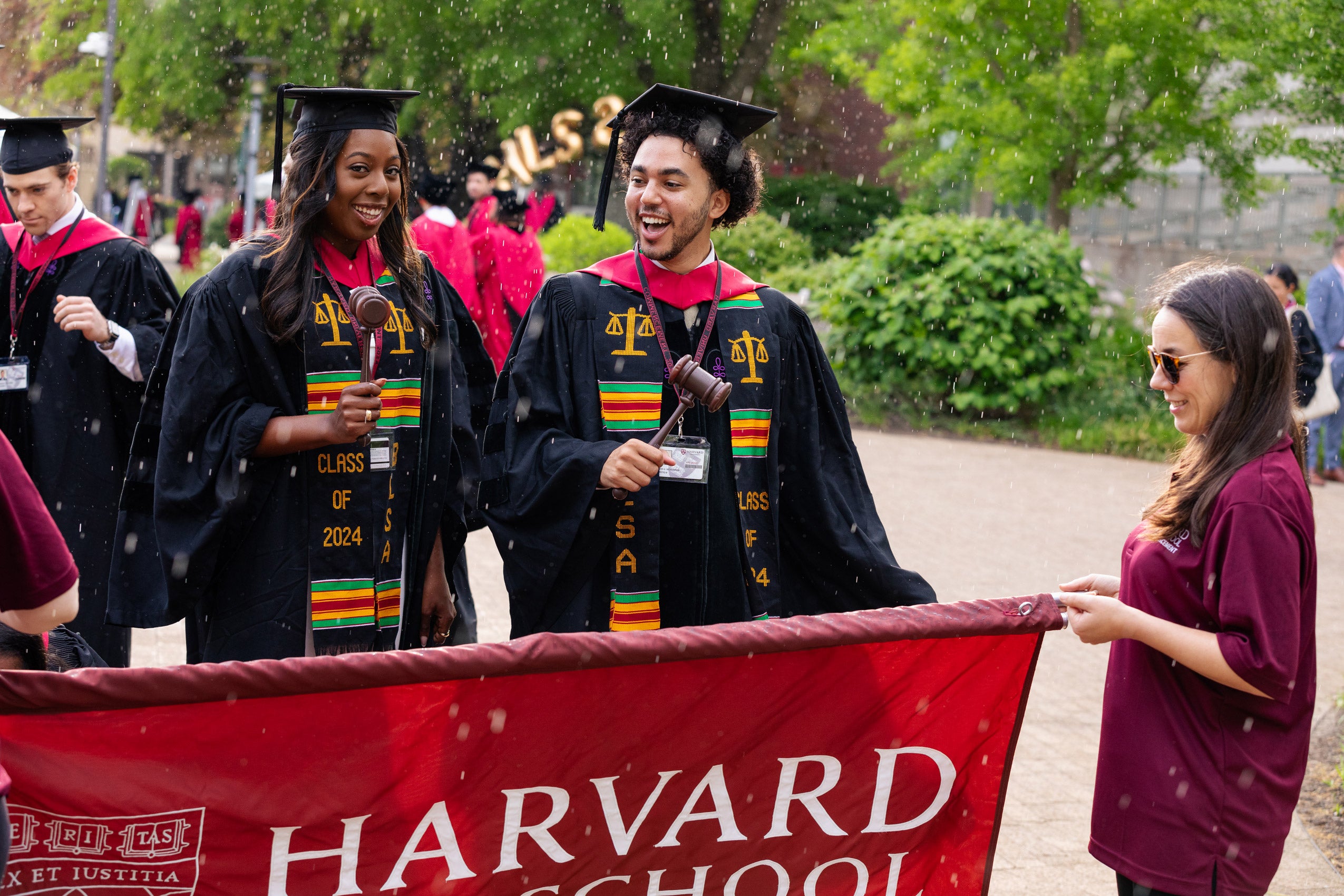 Students in cap and gown stand behind a Harvard Law School banner in preparation for a procession as it starts to rain