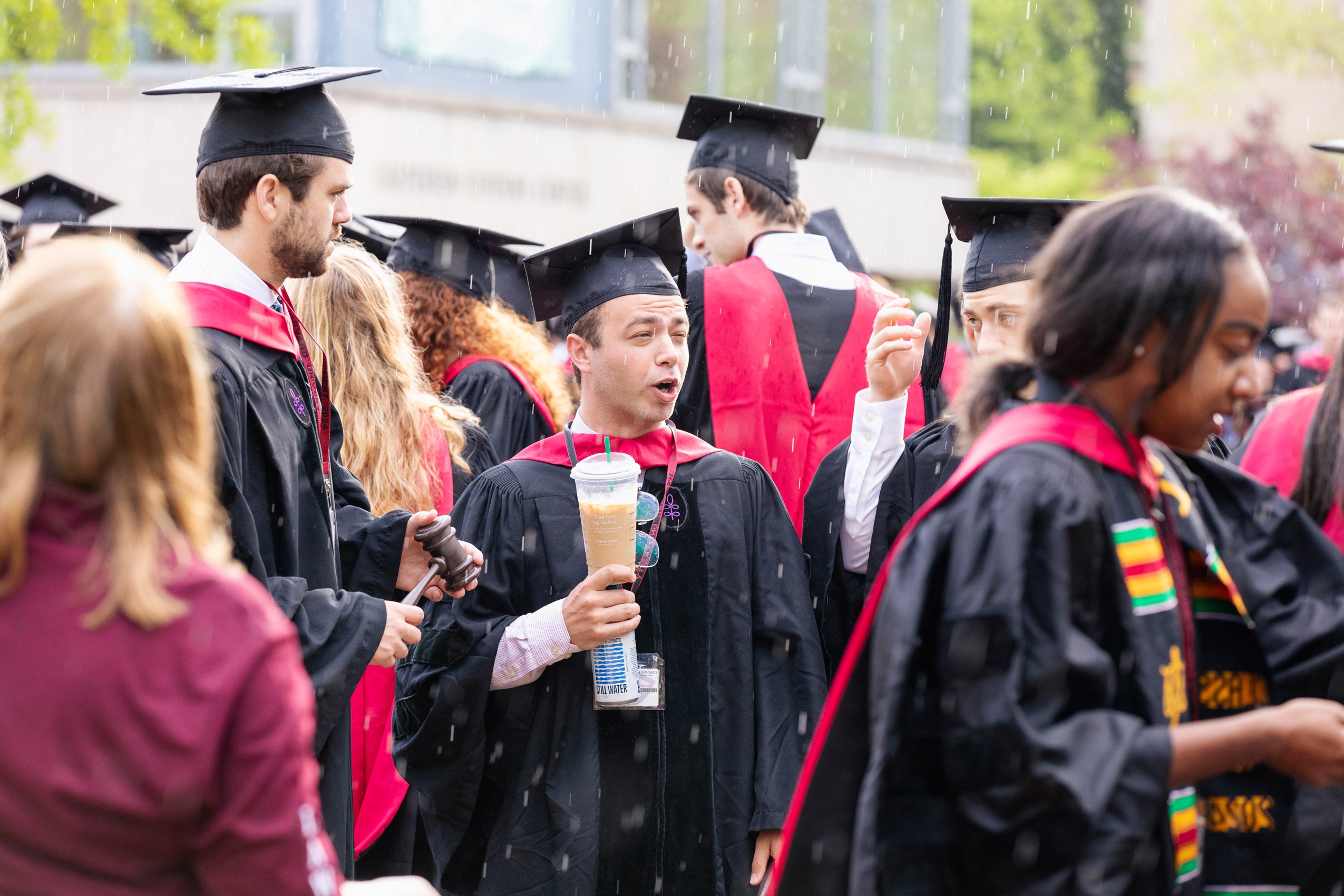 A student in cap and gown in a crowd of others holds a cut of coffee as it starts raining