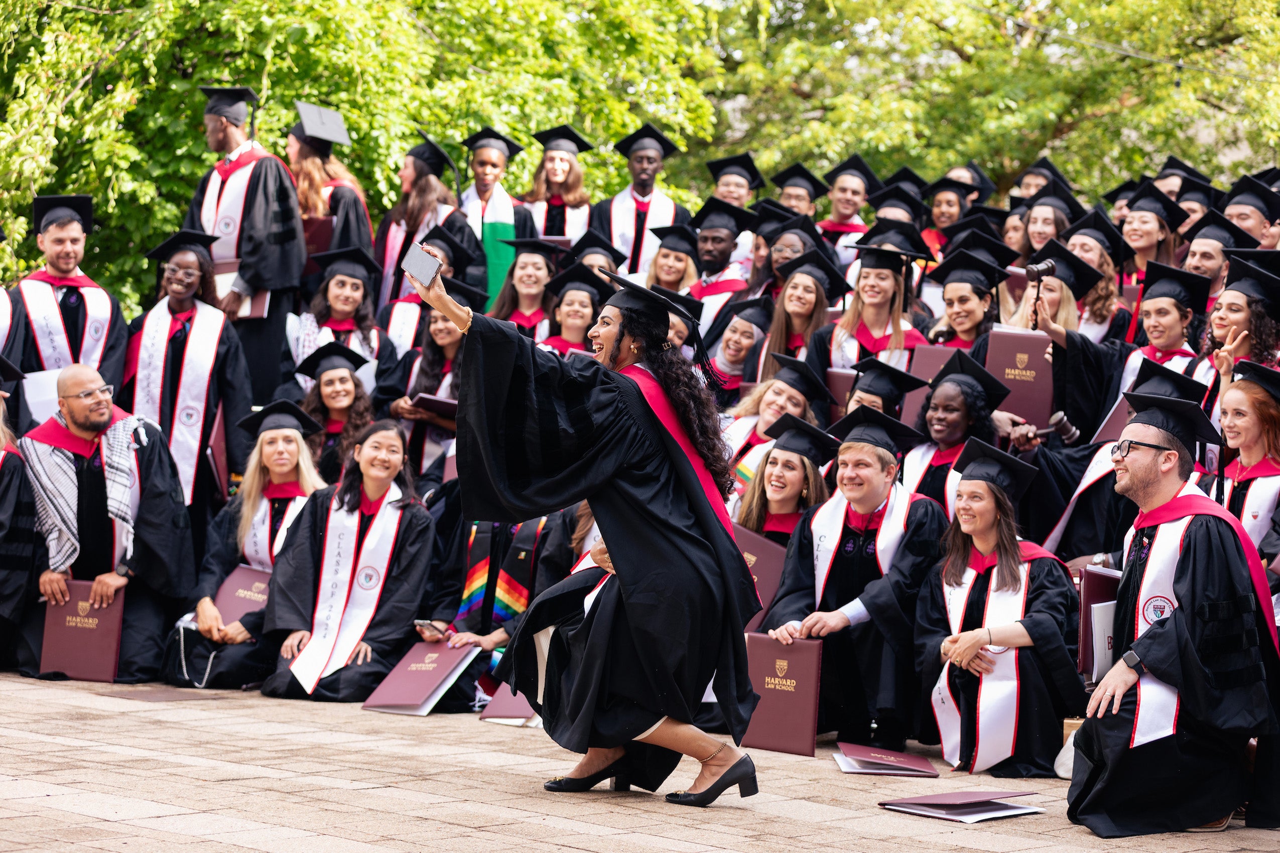 A woman in cap and gown takes of selfie in front of a group a group of other graduates posing for a group shot.