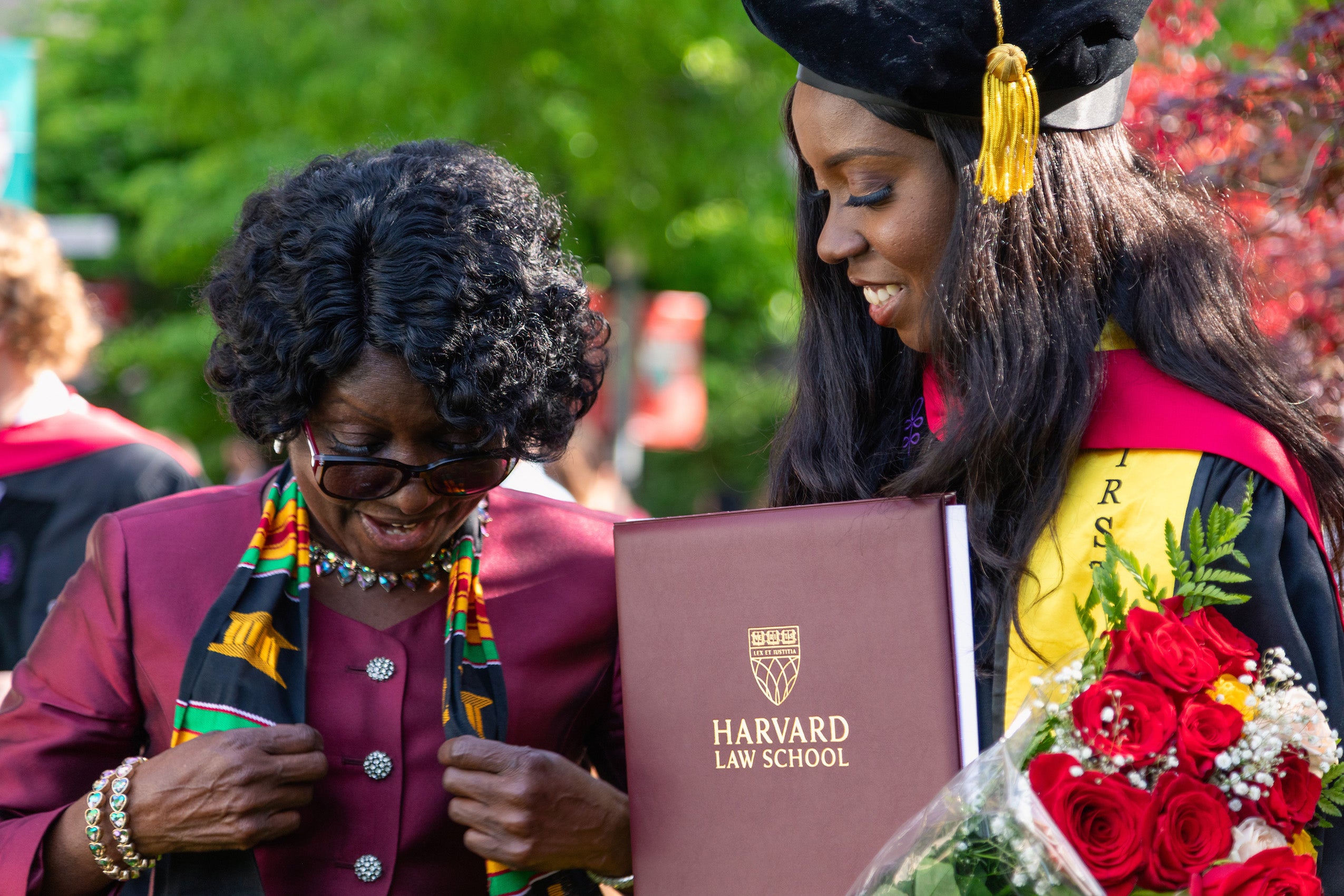 A woman in cap and gown holding a bouquet of roses and her diploma looks on as another woman puts on her sash.