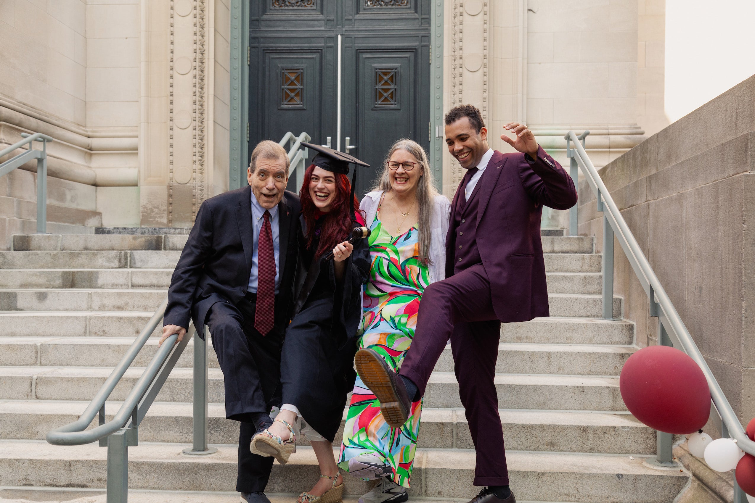 Four people standing near the steps of a building--one in cap and gown kick up their heels in a fun moment.