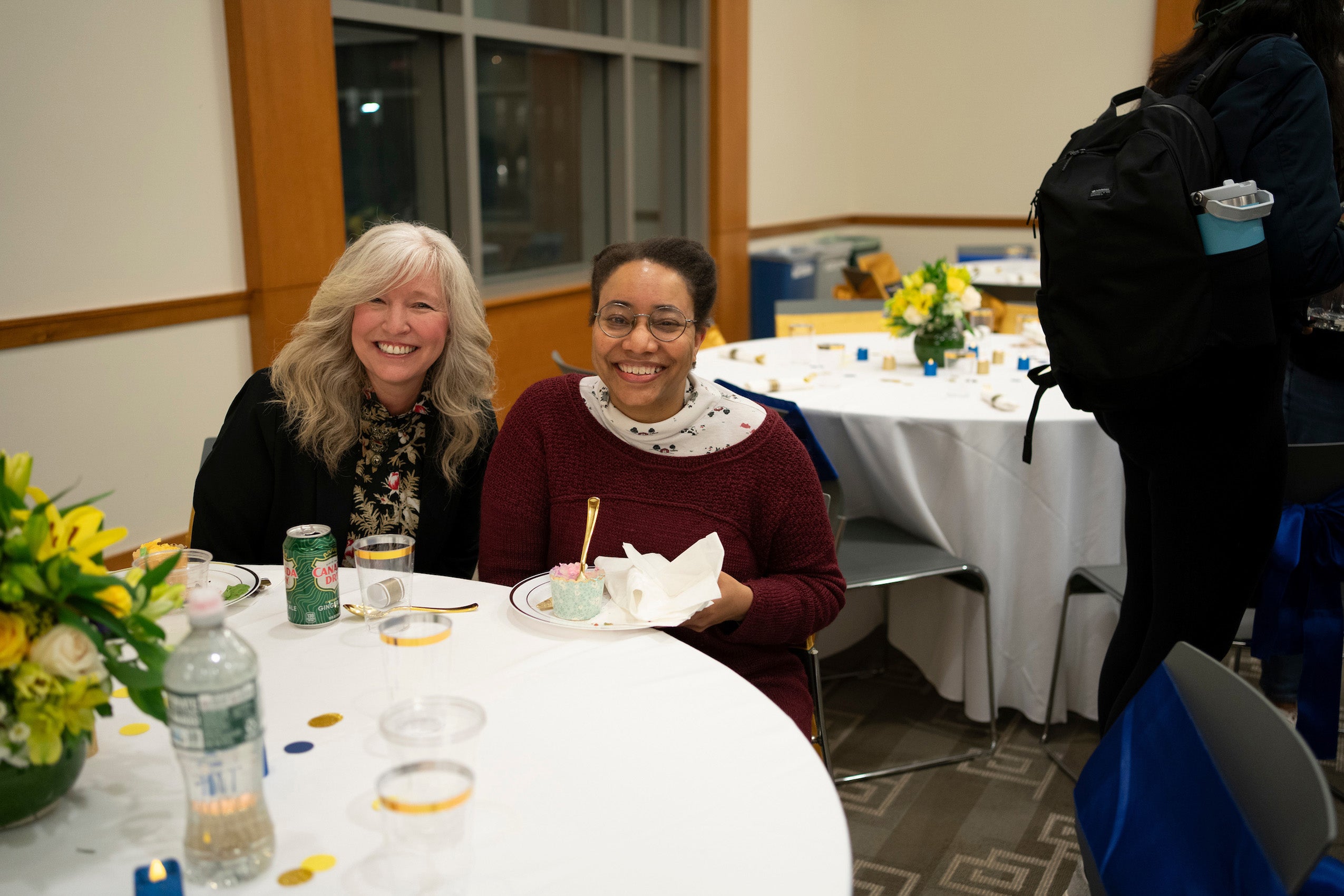 Two woman sitting at a table pose for the camera at an event