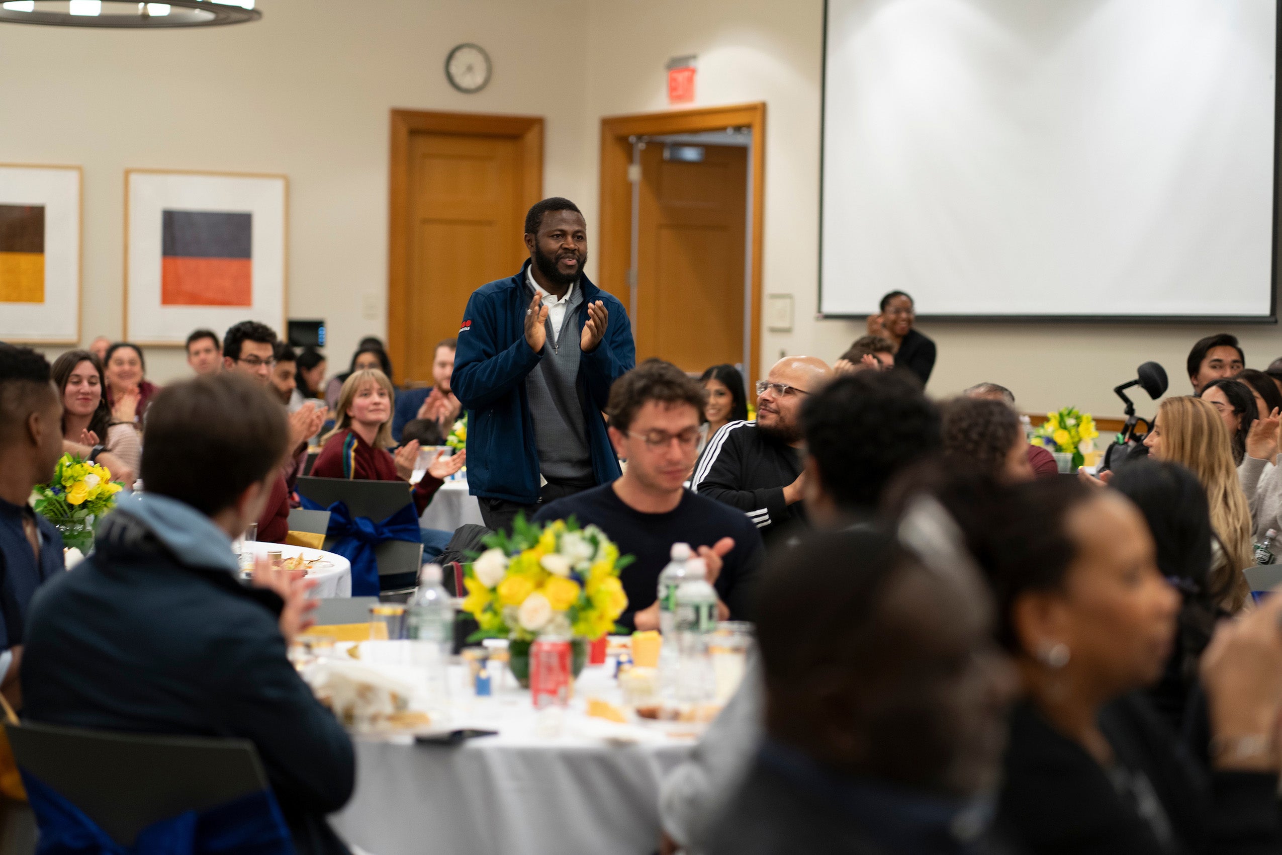 Man standing up clapping in a room filled with people sitting at tables attending an event.