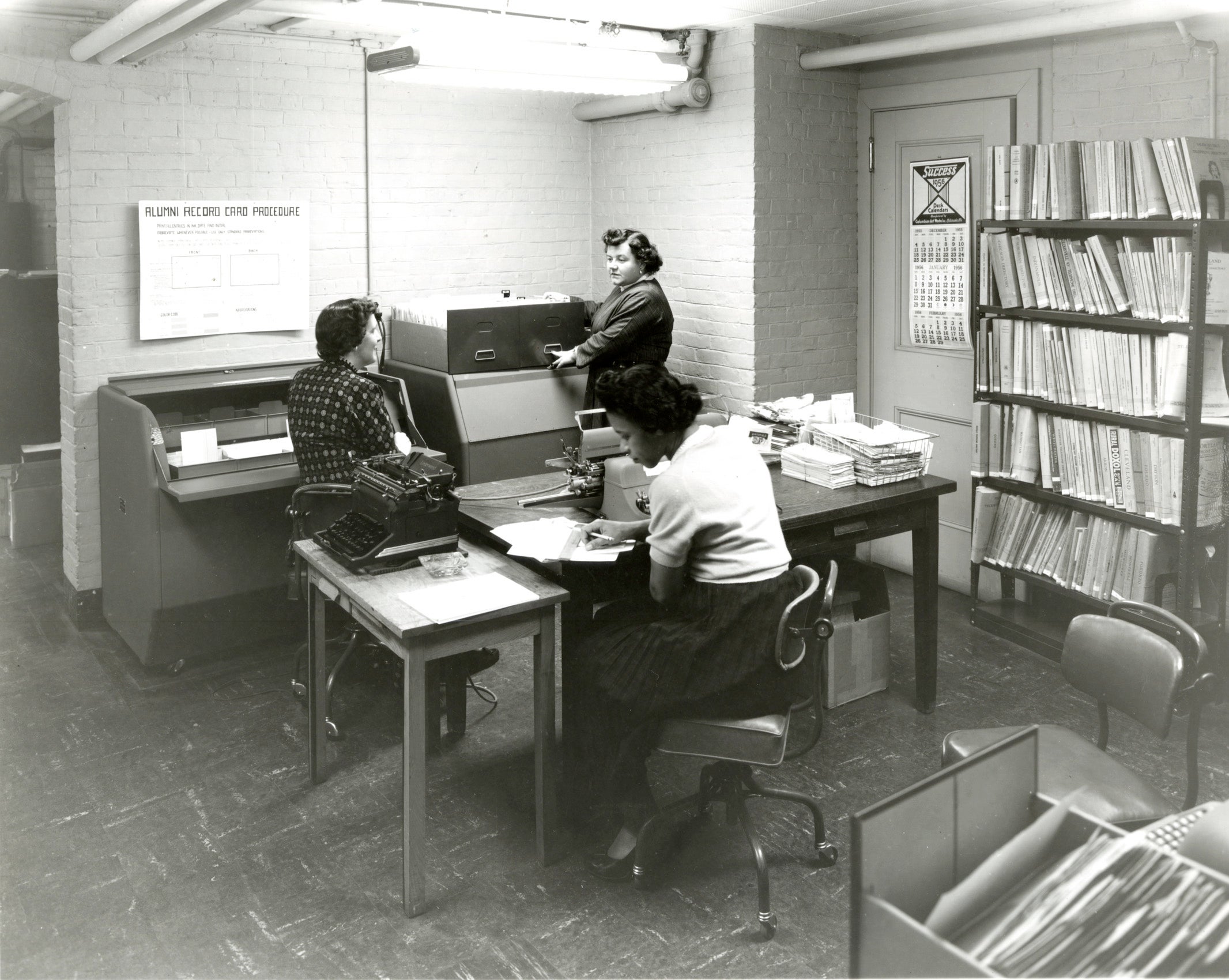 Three woman working in an office with an electronically rotated space saving filing system for alumni addresses.