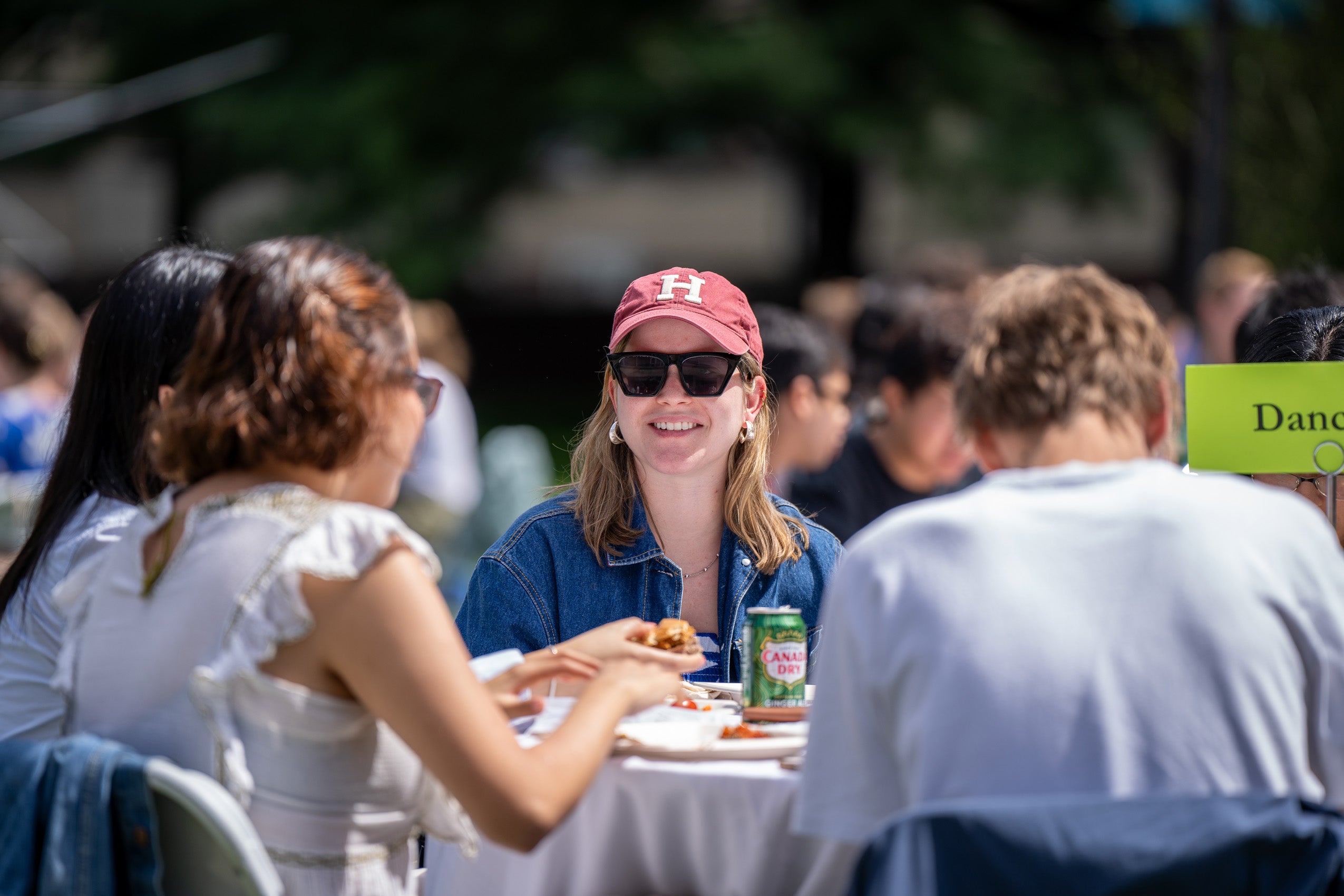 A student wearing at Harvard hat looks up from round table of other students at a table has a sign
