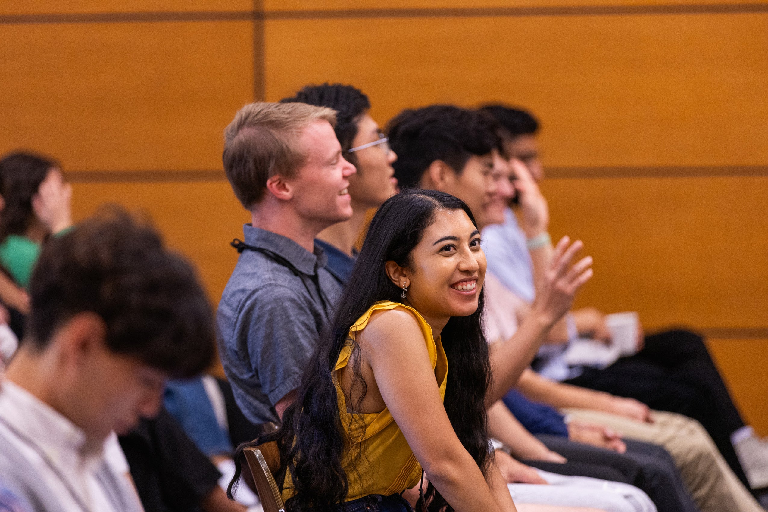 A women in a row of students at an event smiles as she listens to remarks.