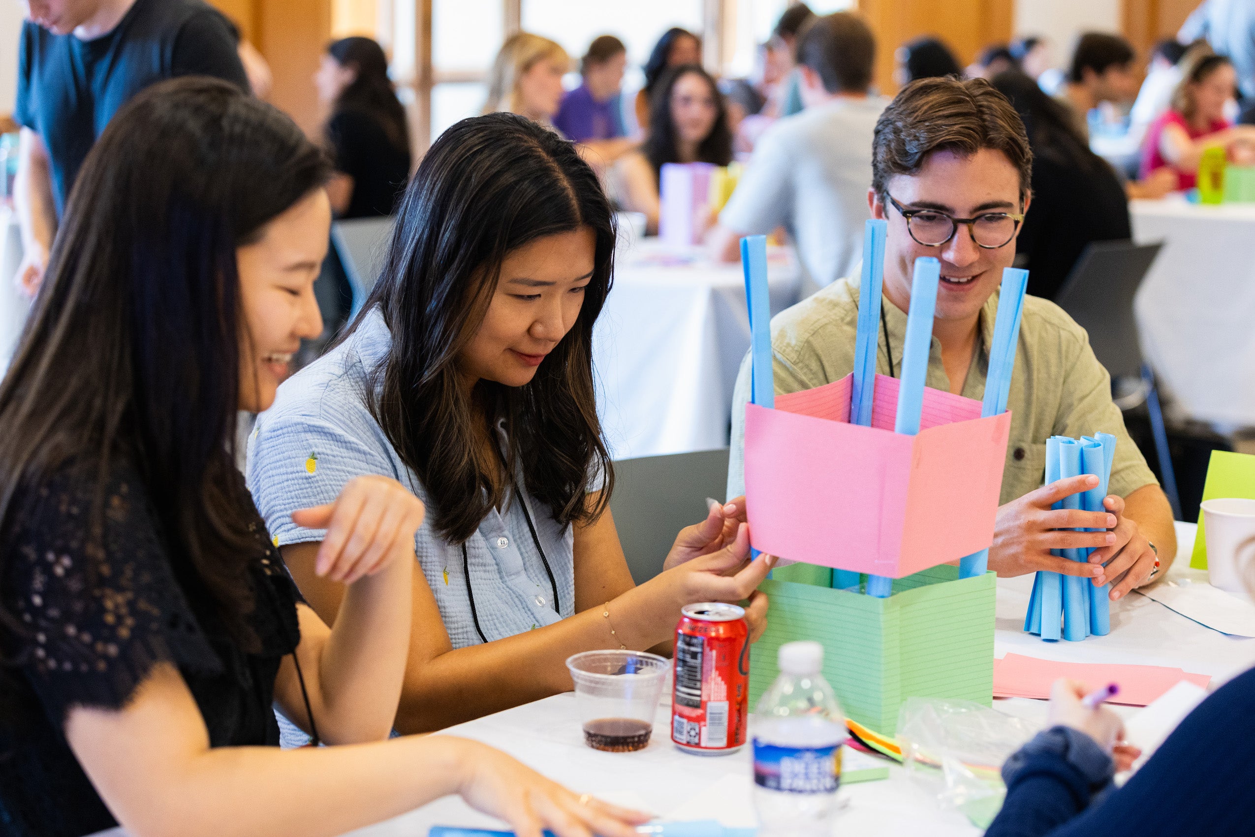 Three people sitting at a table building a paper structure as a team.