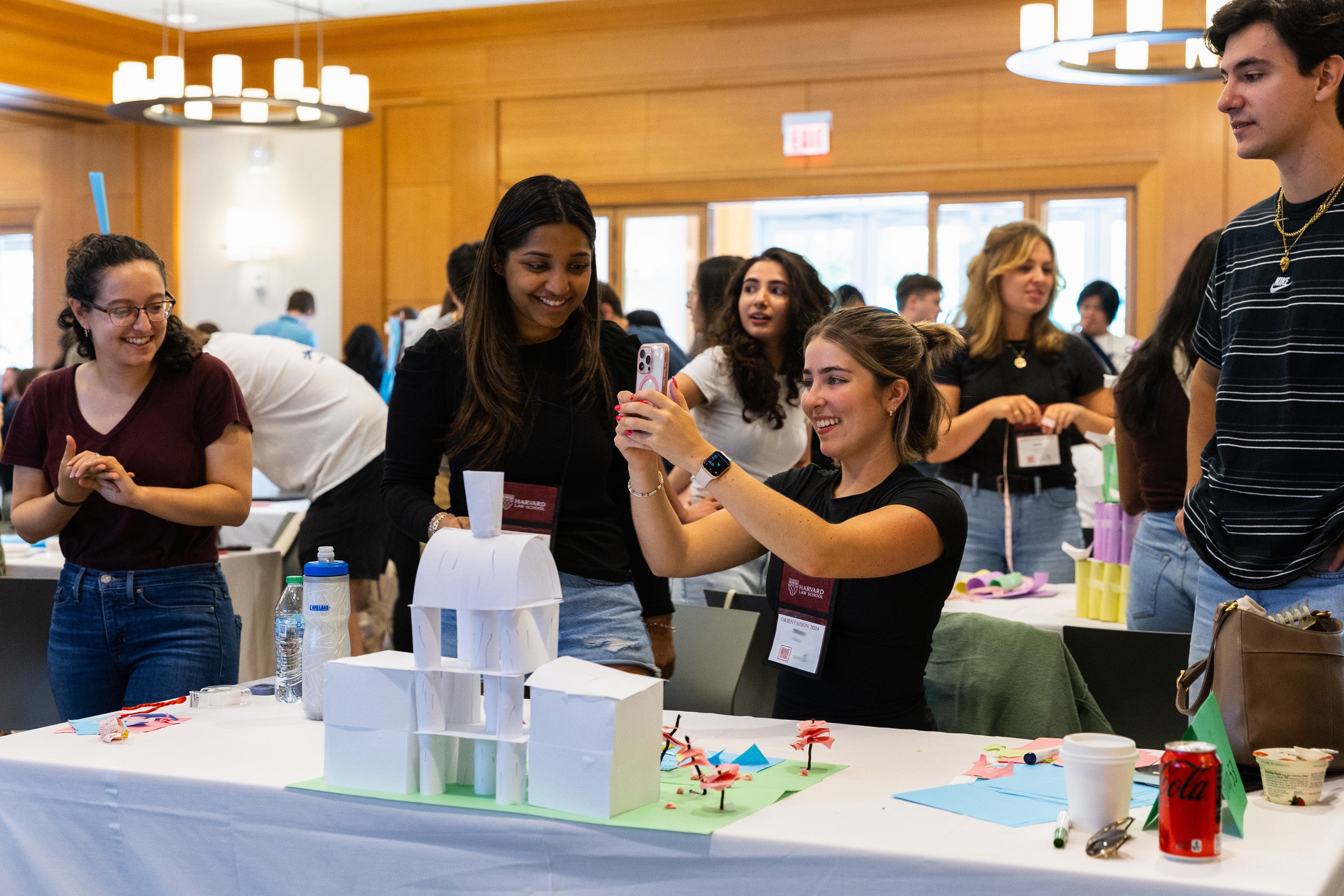 A student sitting at a table takes a photo of a paper structure she and her team members built during a teamwork session during orientation.
