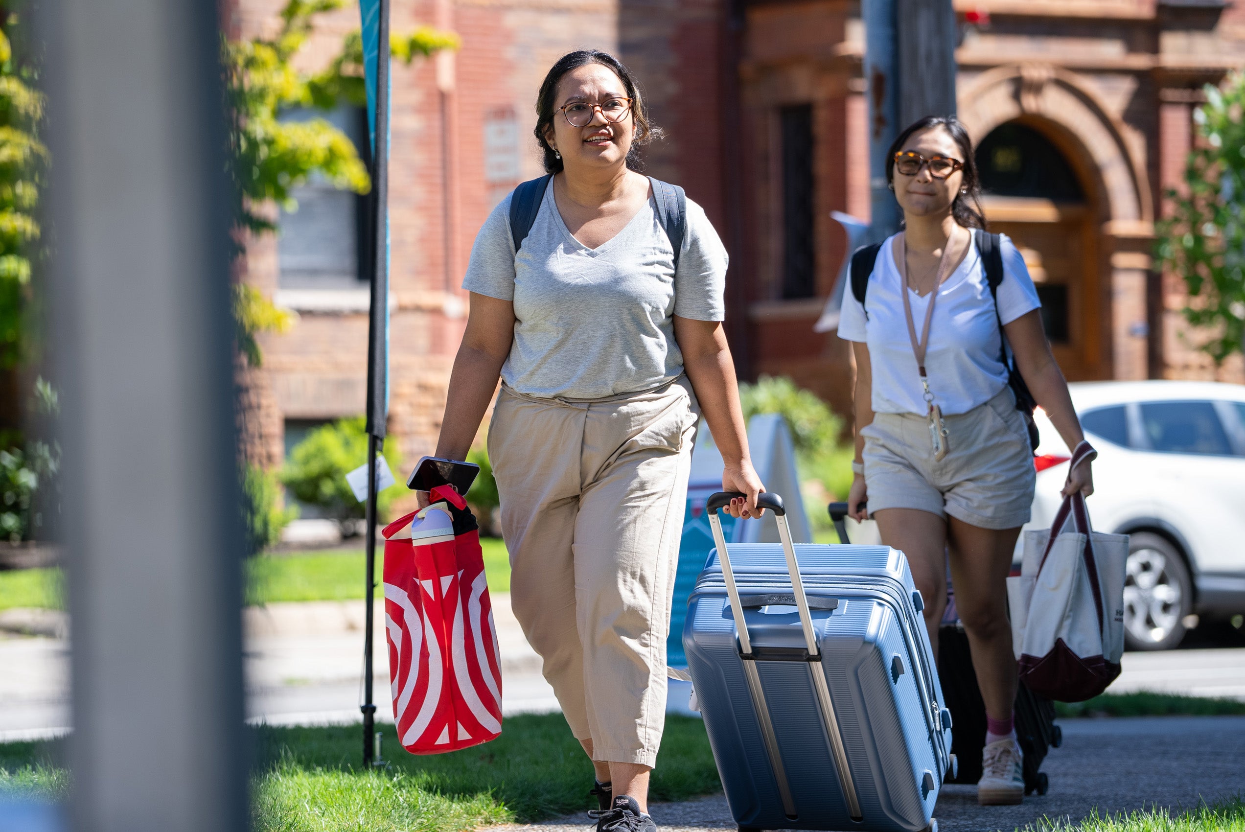 Two women lugging suitcases and bags walking on campus on move in day.