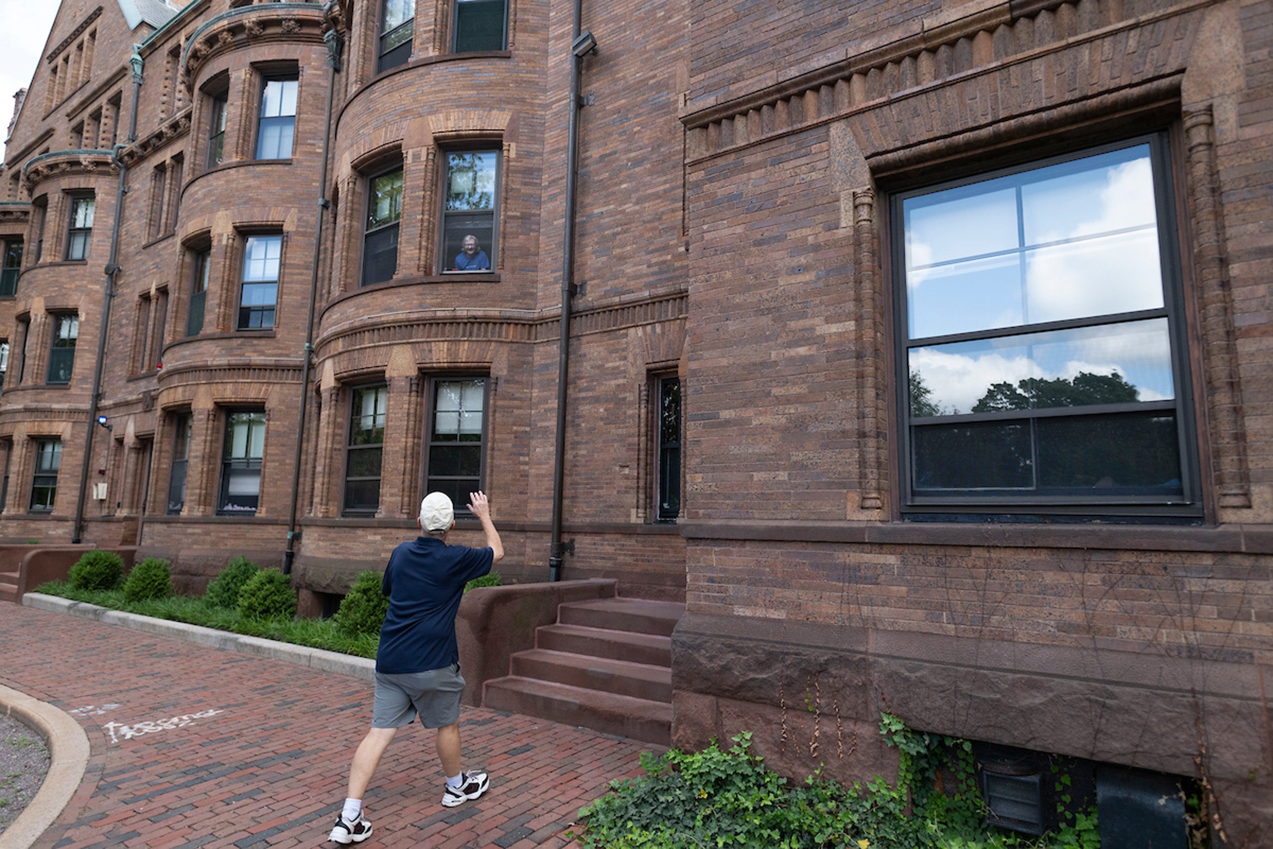 A man waves to someone looking out of a window of a brick dorm on move in day