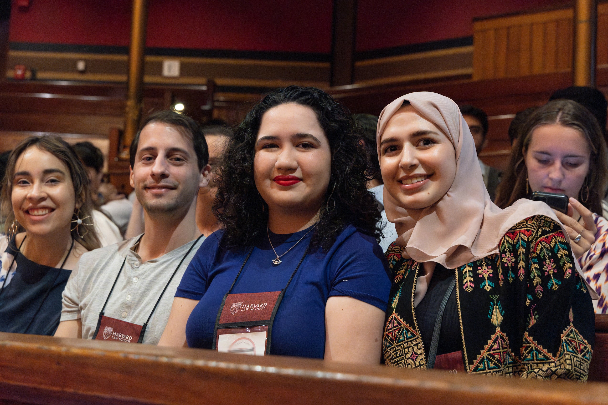 Four students sitting in a row at Sanders Theater pose for the camera before the dean's remarks.