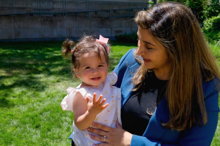 A woman in a blue jacket holds a smiling baby in a white dress.