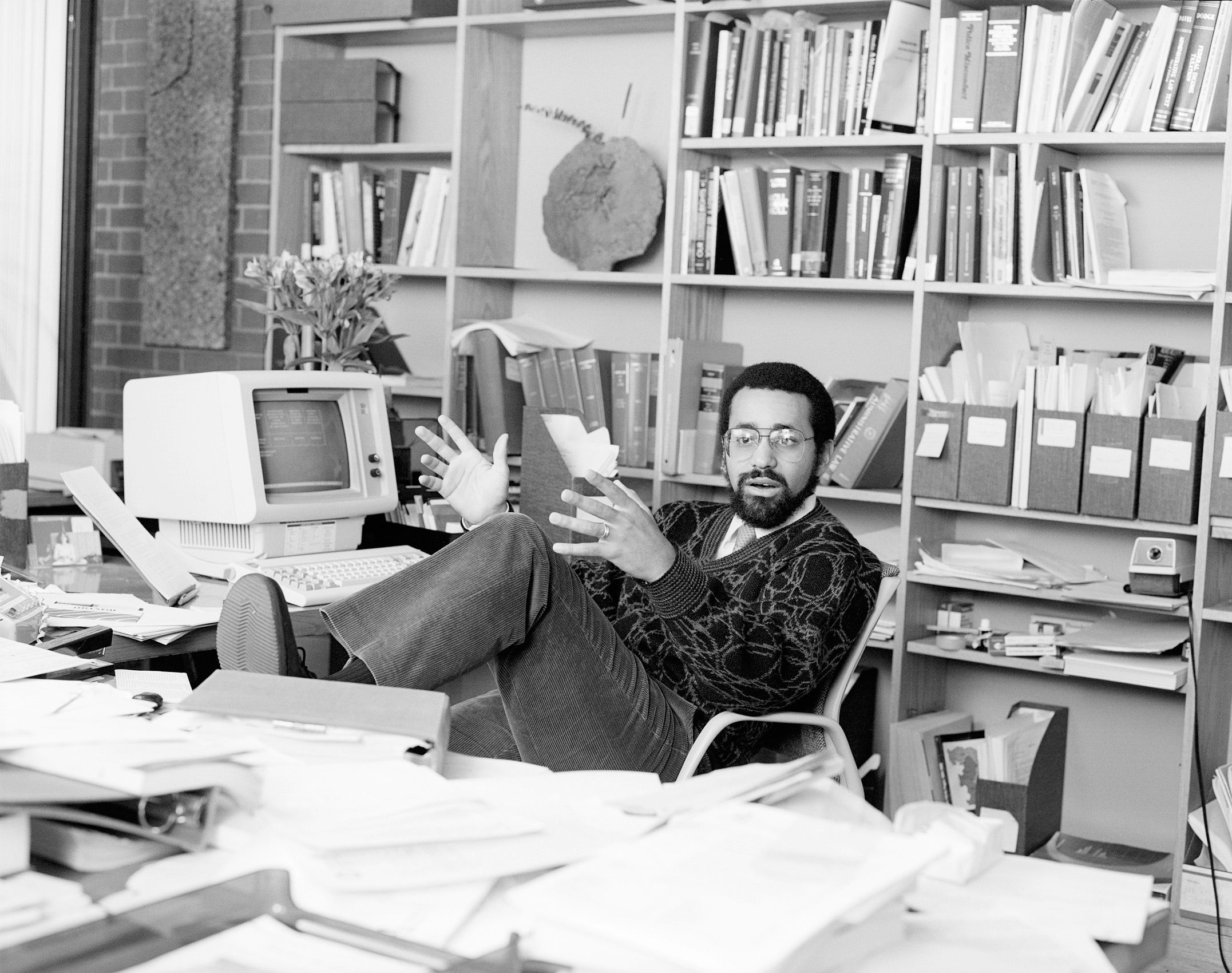 A portrait of Christopher Edley in his office sitting at a desk in front of a wall of bookshelves filled with papers and books.