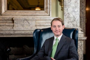 A portrait of Michael Adams sitting in a tall leather care in front of a marble fireplace.