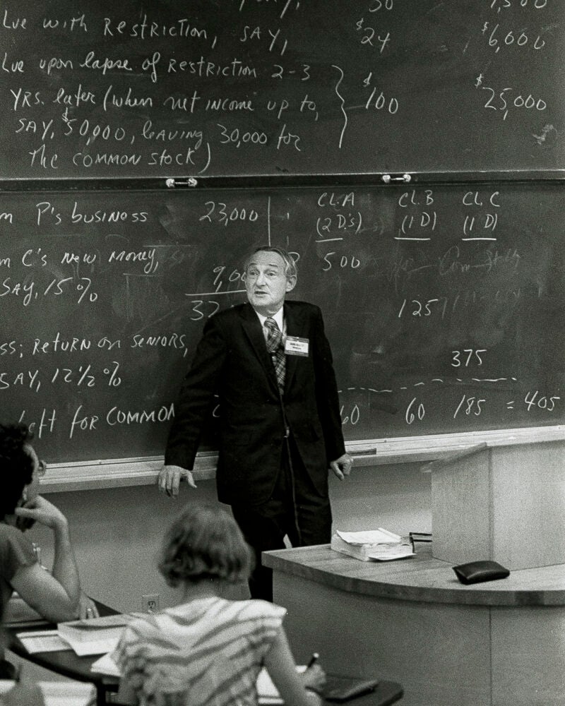A portrait of a man leaning against a blackboard covered in information in a classroom