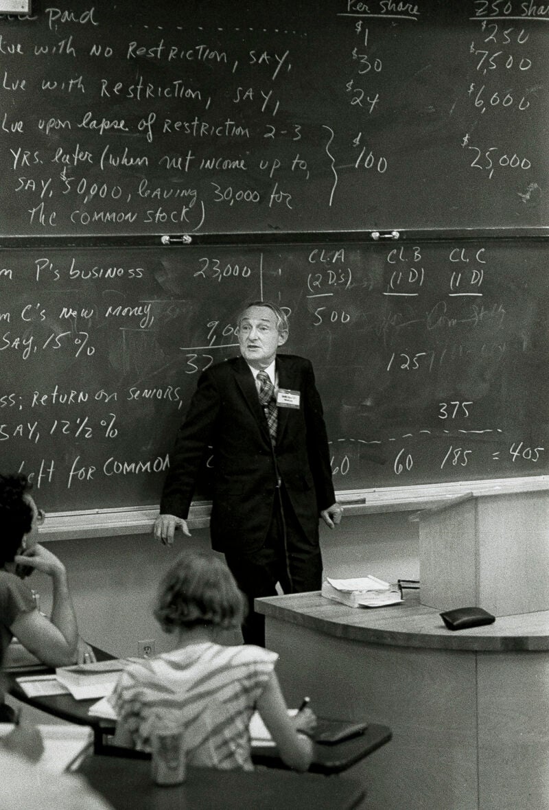 A portrait of a man leaning against a blackboard covered in information in a classroom
