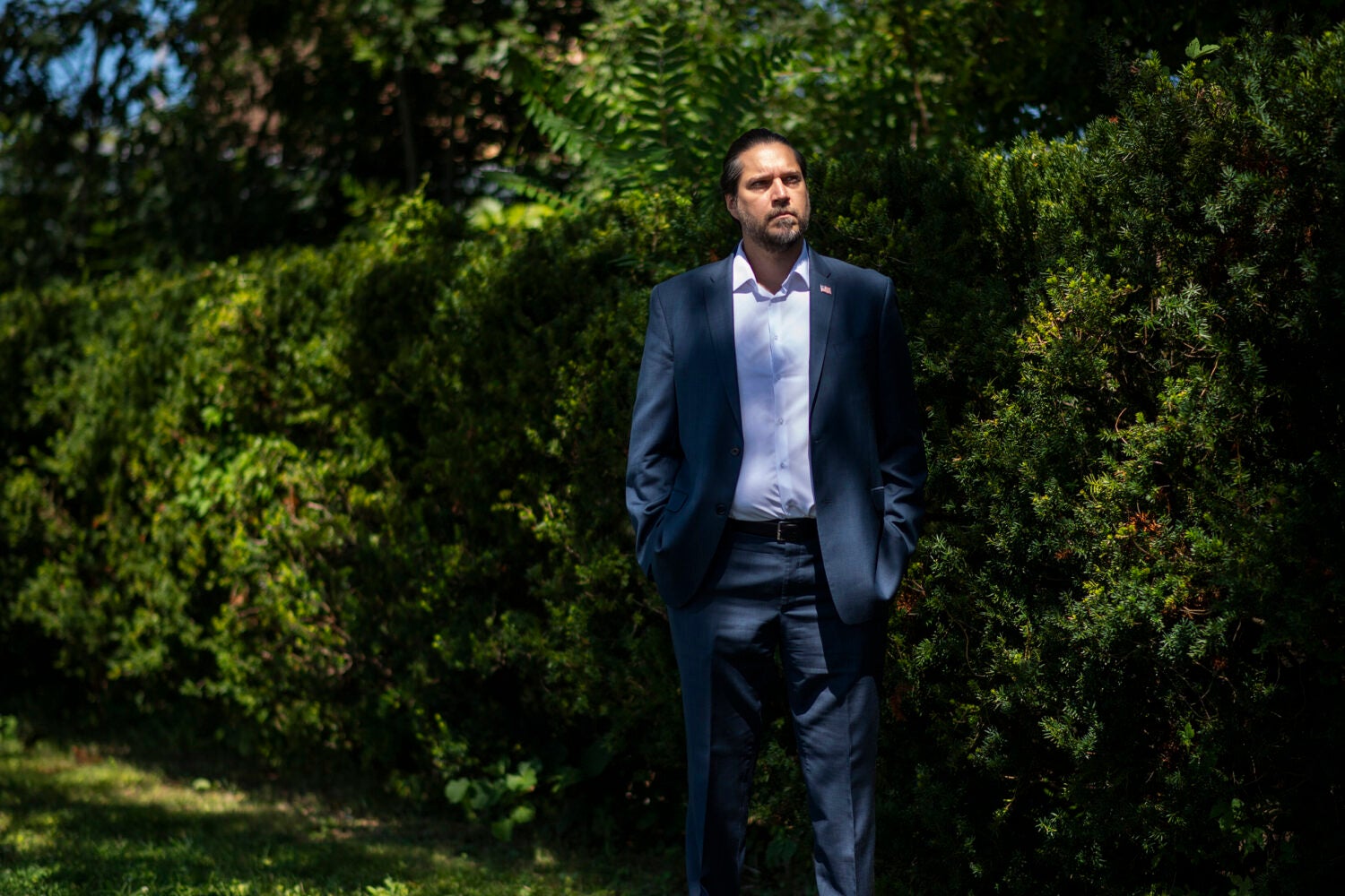 A portrait of a man in a casual suit standing in front of a row of bushes.