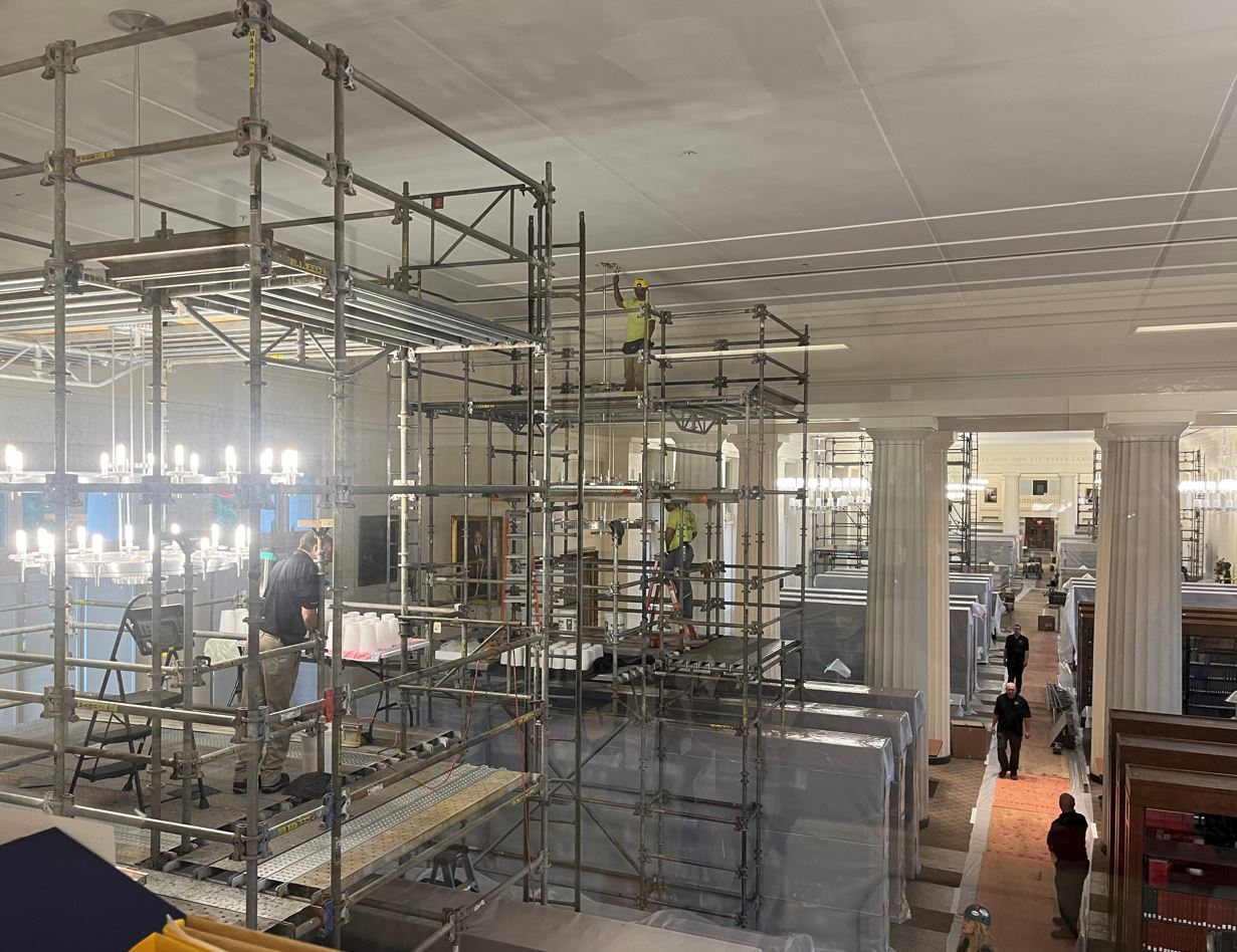 The Reading Room in Langdell Hall with several scaffold and people on them repairing chandeliers, bookcases covered with plastic, and people walking nearby.