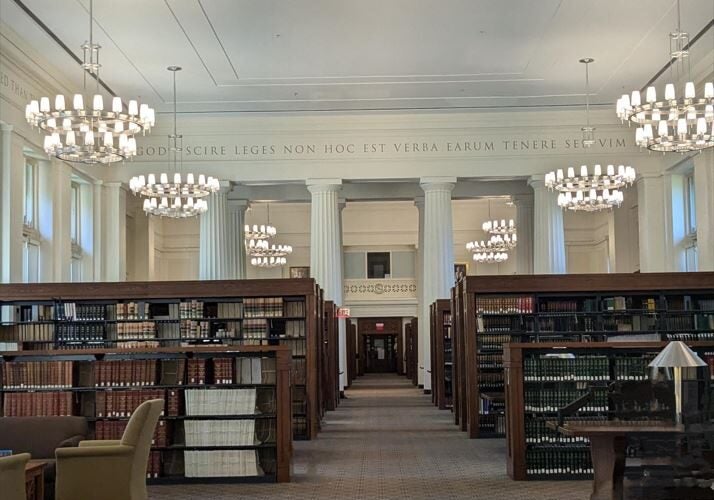 An expansive room with Greek Doric columns, tall ceilings, bookcases, and chandeliers.