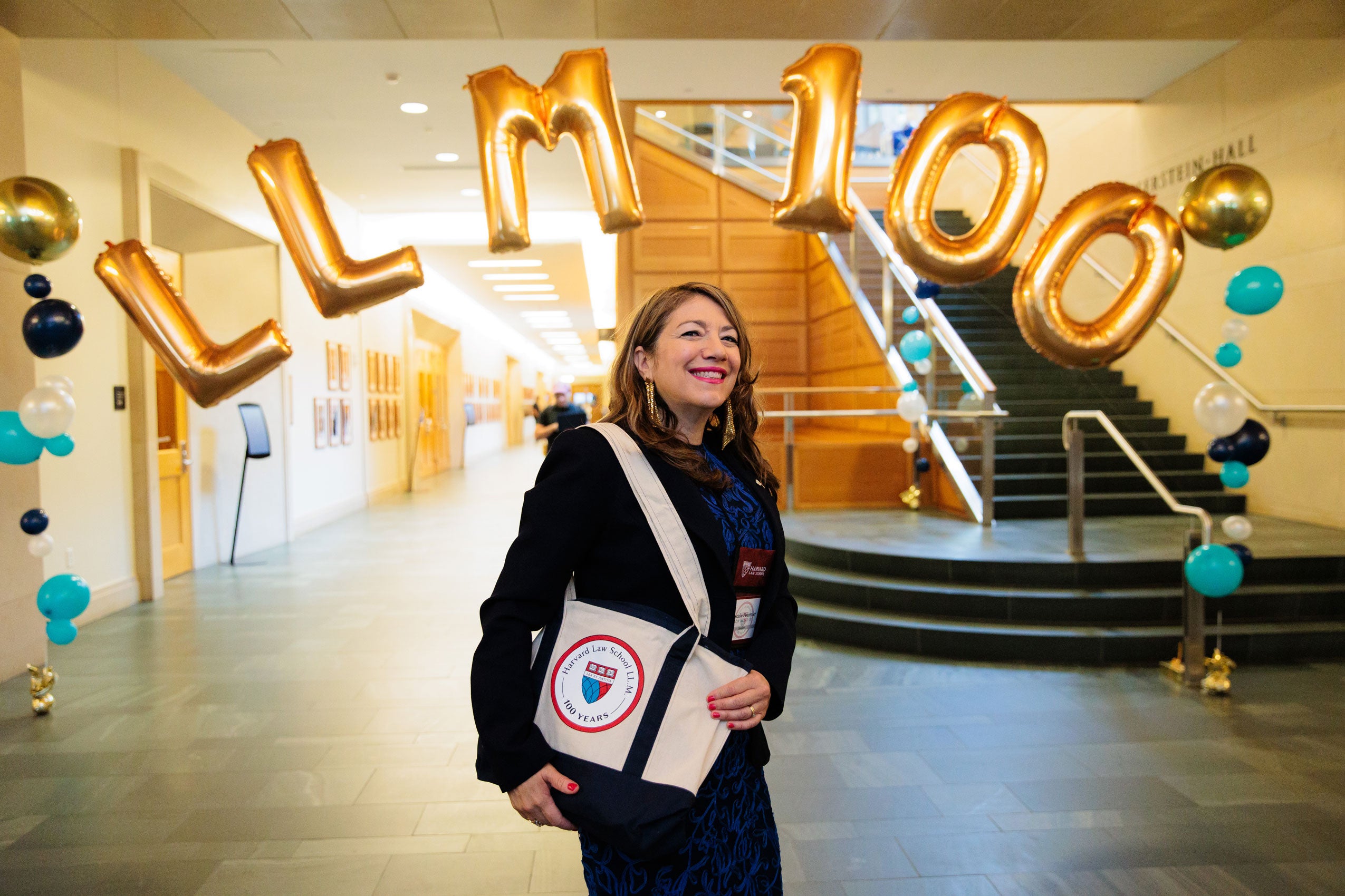A woman stands under gold balloons that spell LL.M. 100 in the hallway of a building.