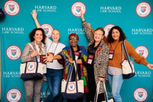 A group of women stand in front of a a teal backdrop posing for a photo at a celebration