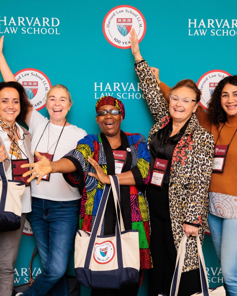 A group of women stand in front of a a teal backdrop posing for a photo at a celebration