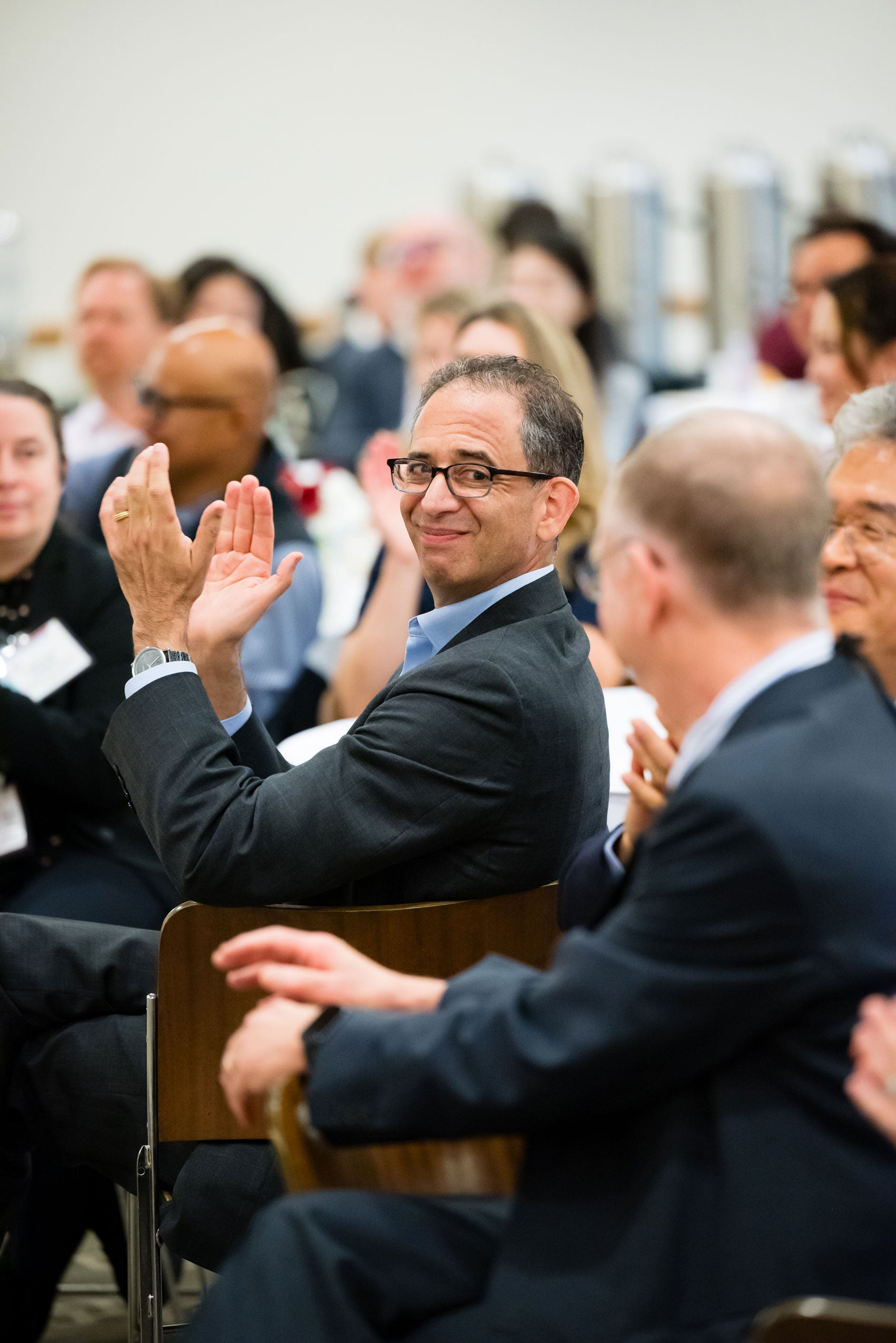 A man claps as he looks over his shoulder in a room filled with people at an event.