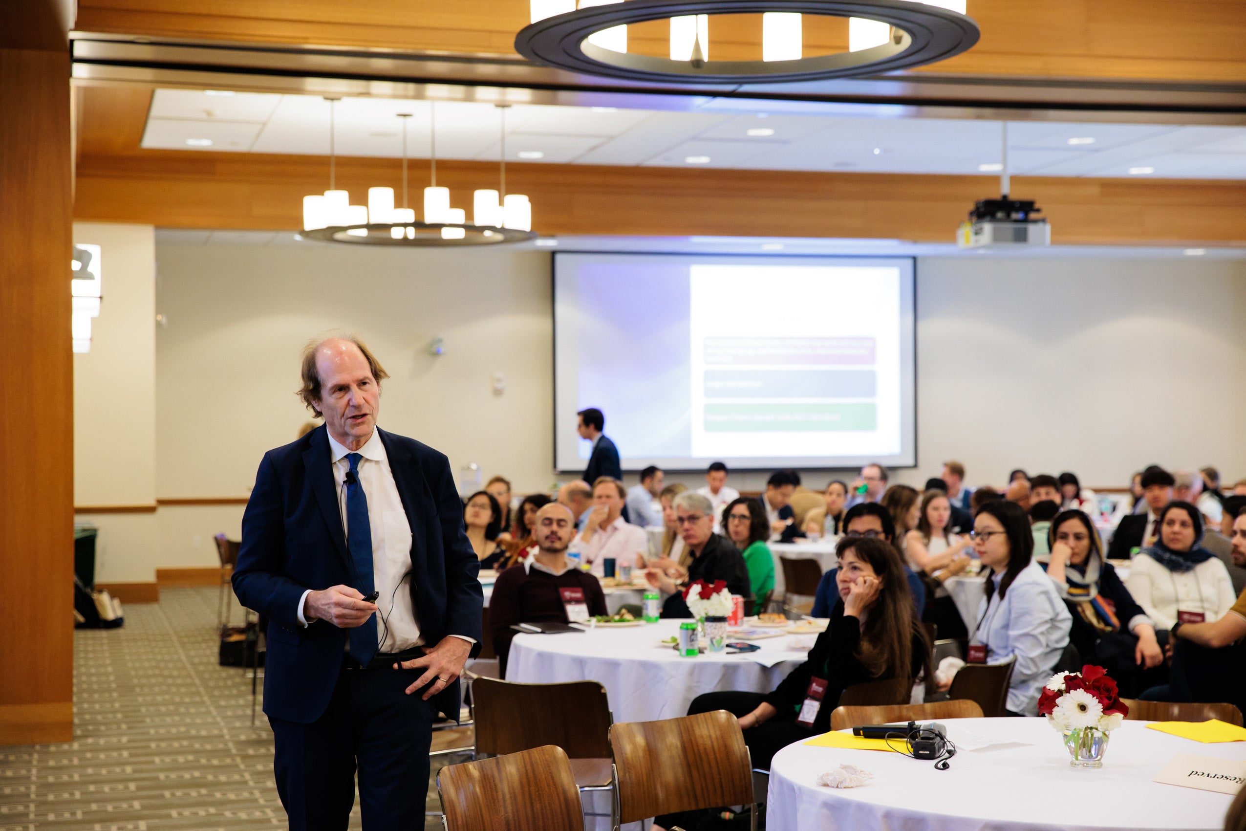A man lectures a large group of people sitting around tables in a room