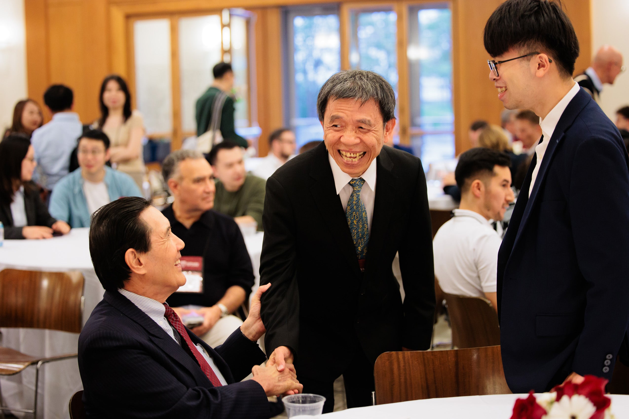 Two men great another at one of the tables in a large room filled with people at tables.