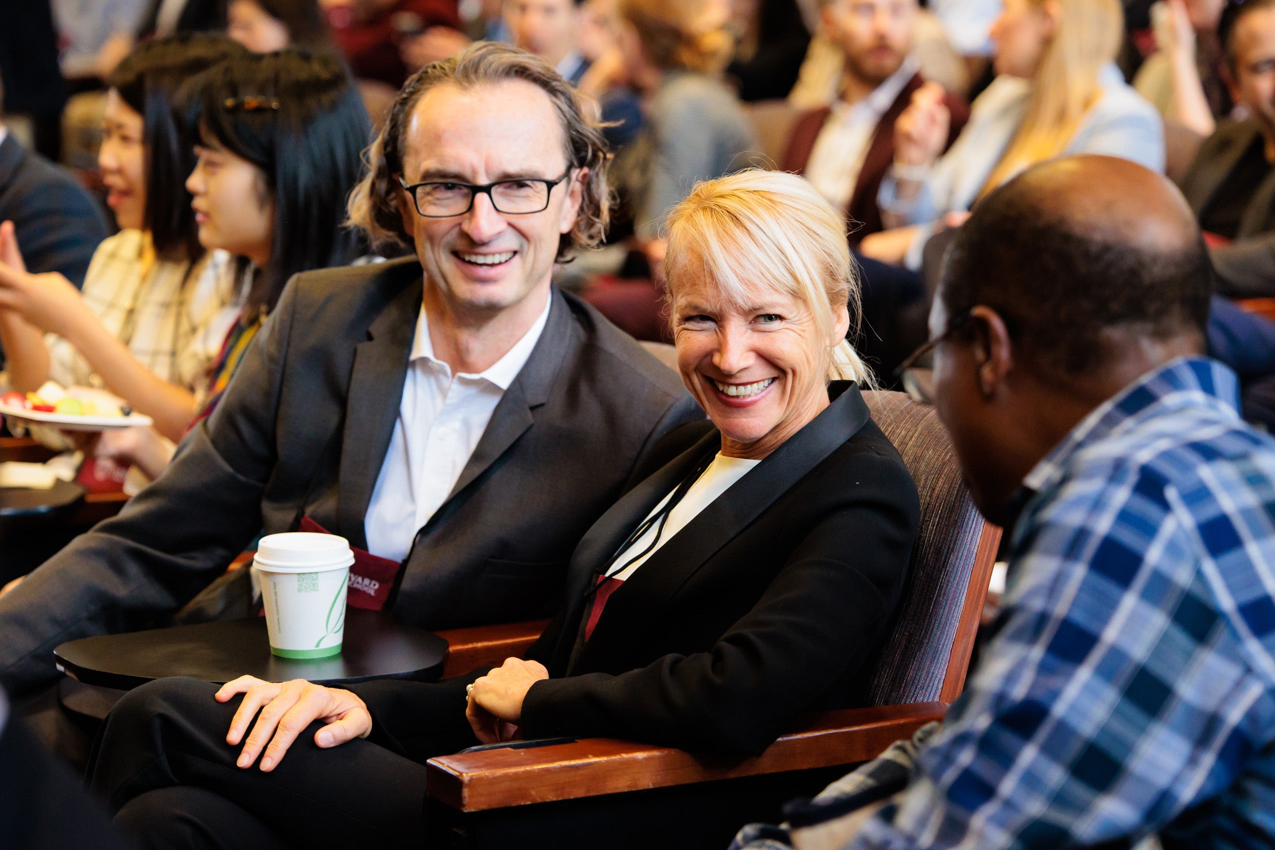 A man and woman sitting in an audience chat with another man sitting nearby