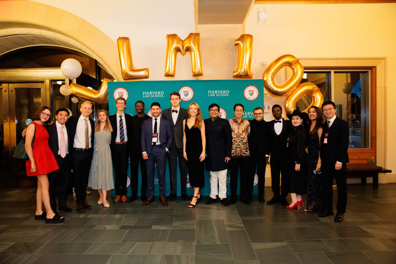 A large group of men and women gather beneath gold balloons which spell LL.M. 100 and in front of a green screen with several 100th anniversary logos