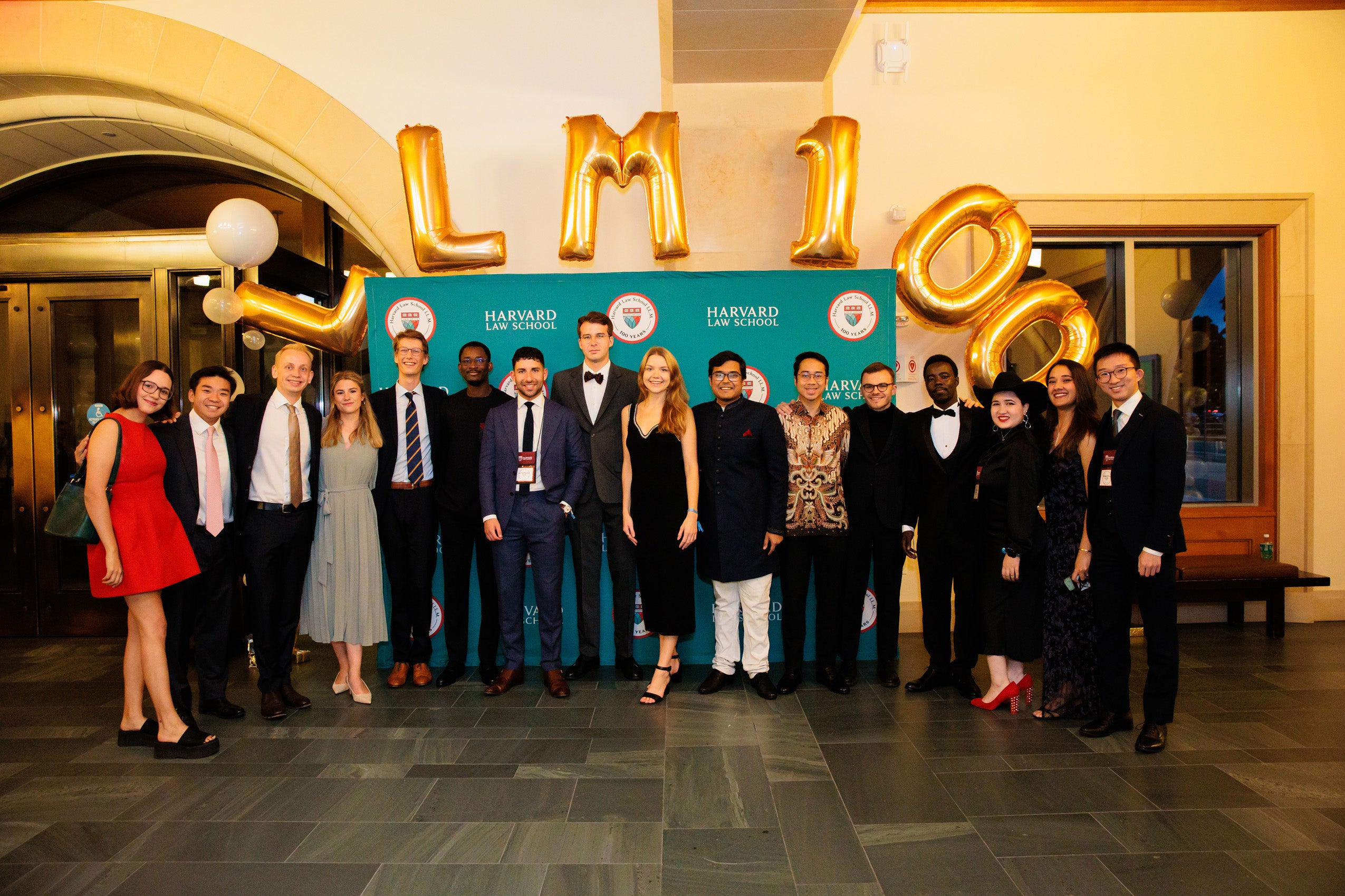 A large group of men and women gather beneath gold balloons which spell LL.M. 100 and in front of a green screen with several 100th anniversary logos