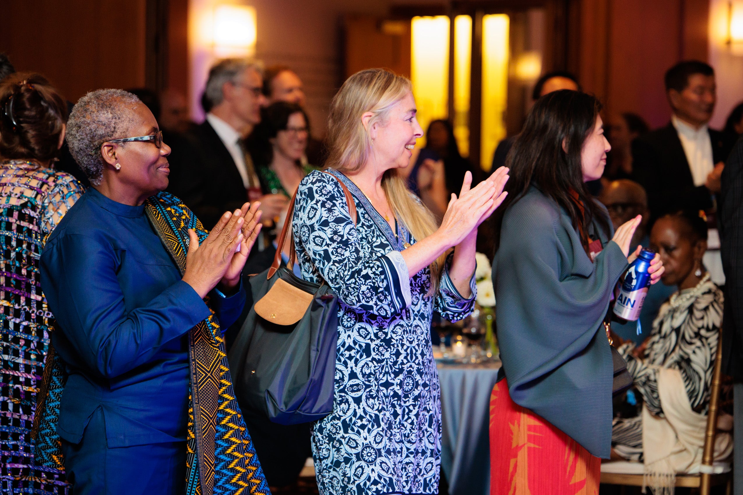 a row of three women clap in a room filled with people at at party