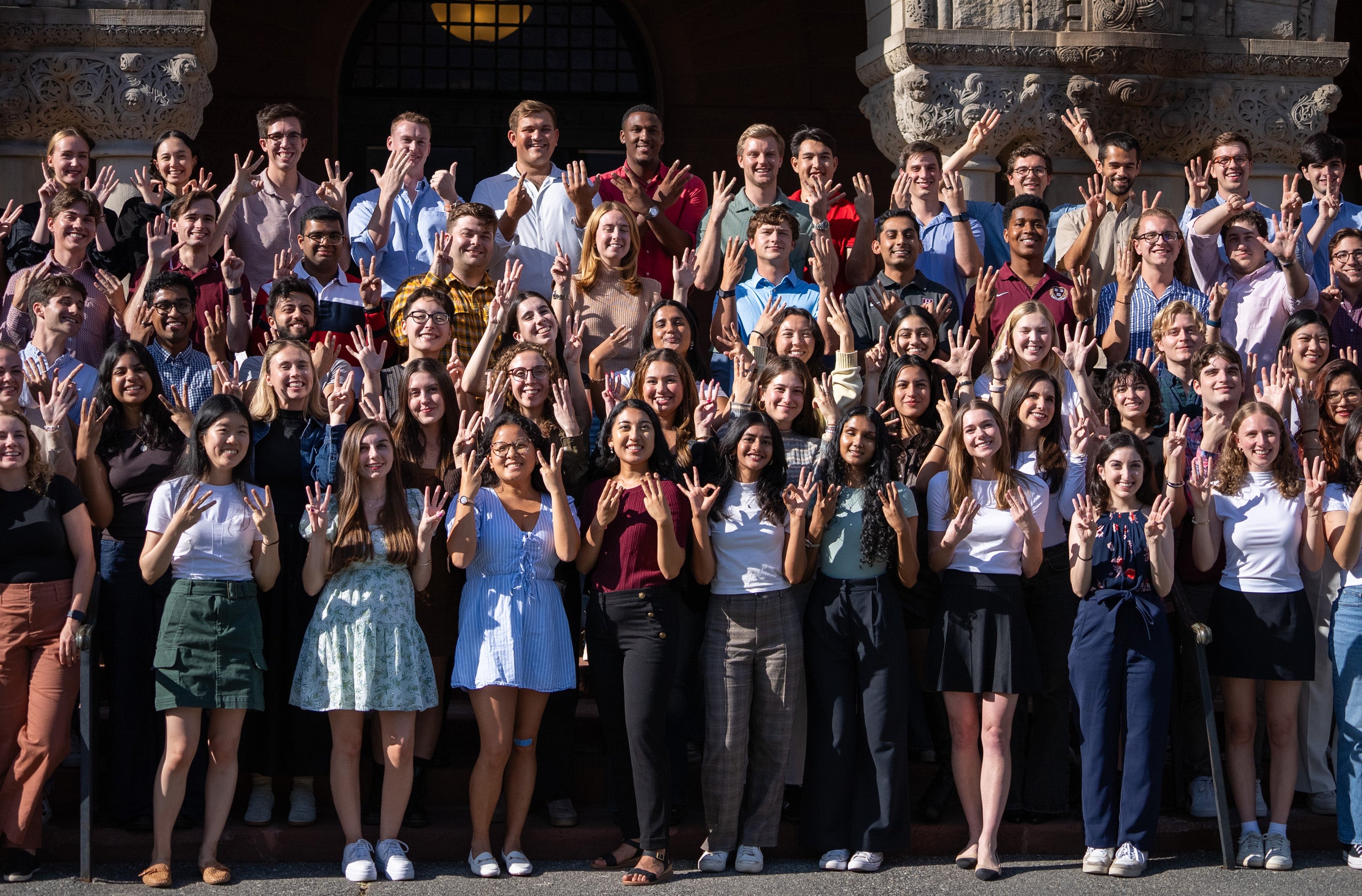 A large group of students in several rows on the steps in front of a building hold up their fingers to indicate their section number in this photo.