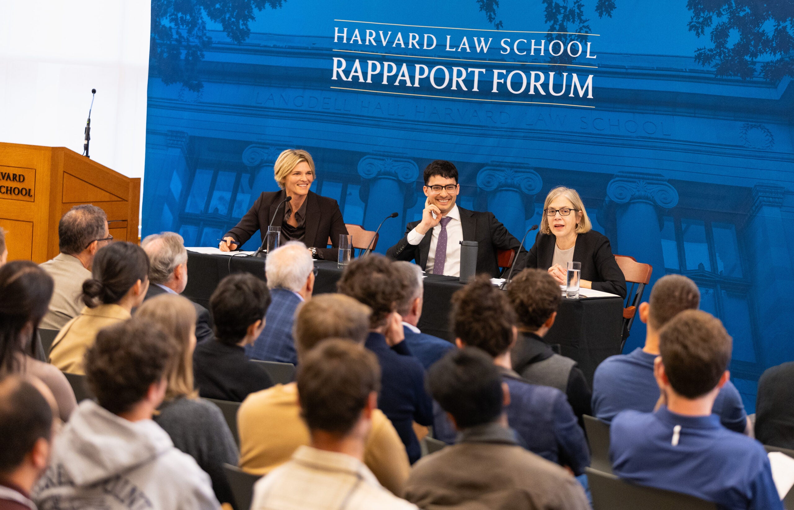 Three people in front of a blue backdrop seated at a table speak to an audience.