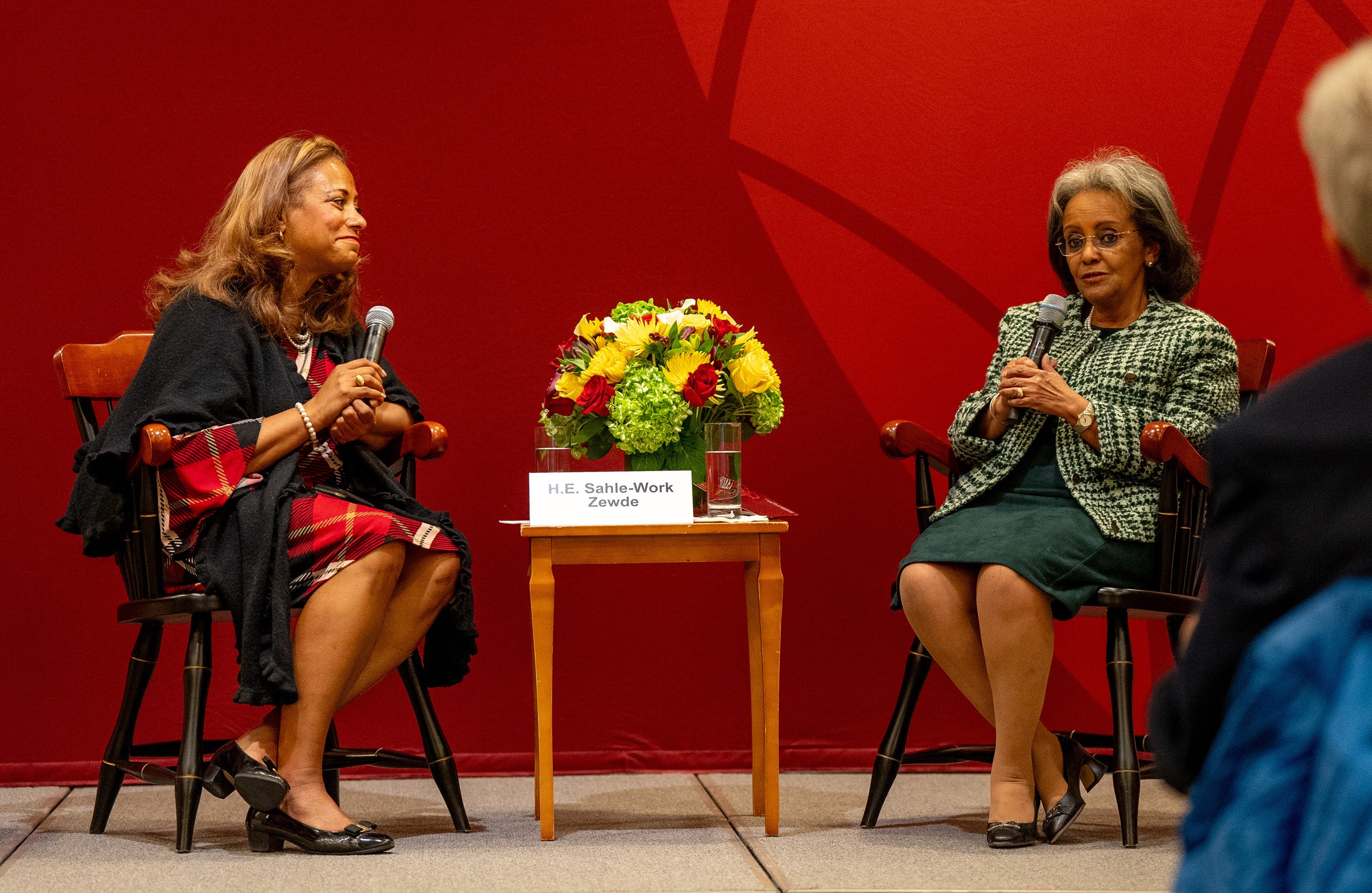 A woman sitting on a stage in front of a crimson backdrop interviews another woman.