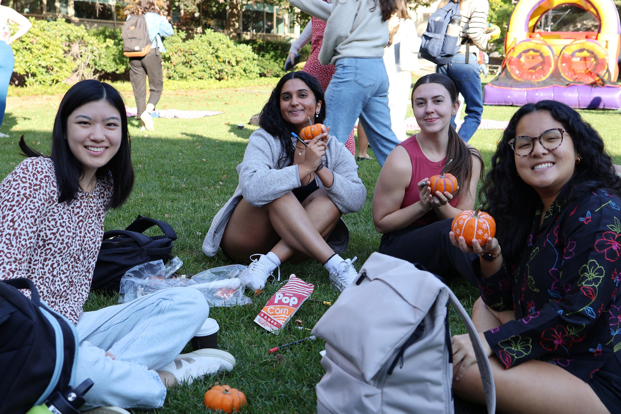 Several students holding painted pumpkins sit on the grass during fall festivities on campus