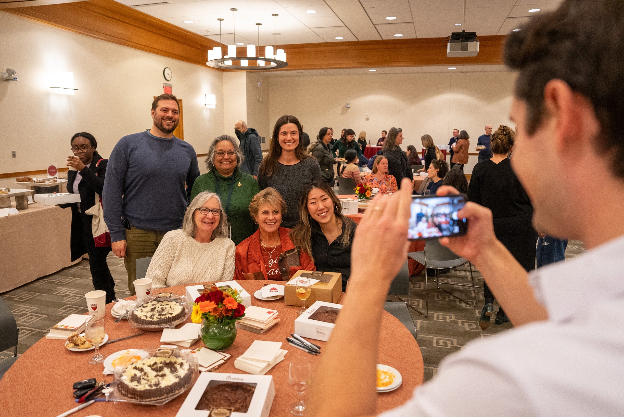 A man takes a photo of a group of people posing behind a table that has some note cards and pies