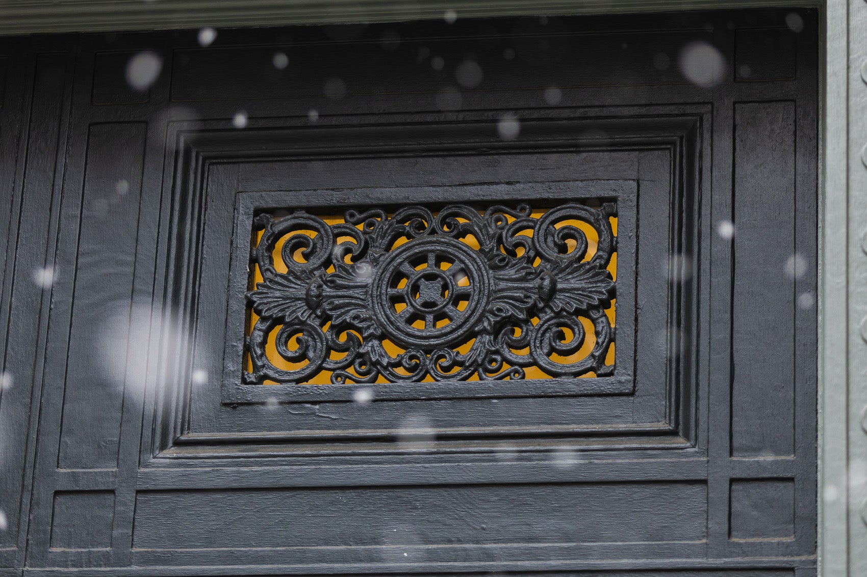 A yellow light shines behind a decorative detail of a black door on a snowy day.