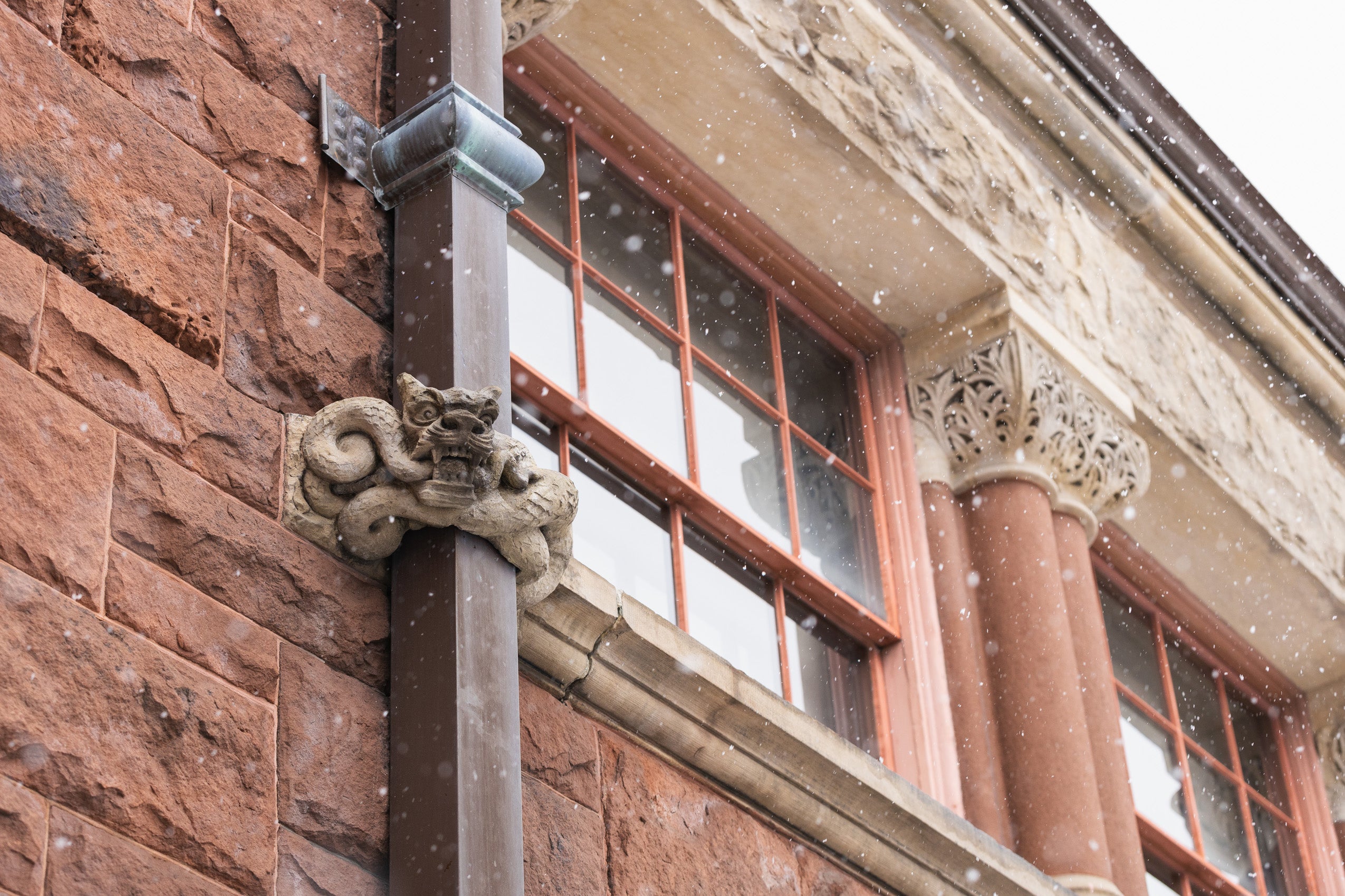 A detail of the top of a red brick building with light colored details on a snowy day. A stone dragon looks lut from a drainpipe.