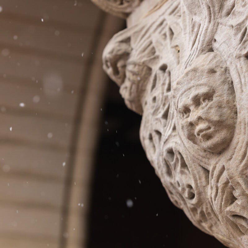 A detail of a carved face figure with a pleasant expression at the top of a stone column on a snowy day.